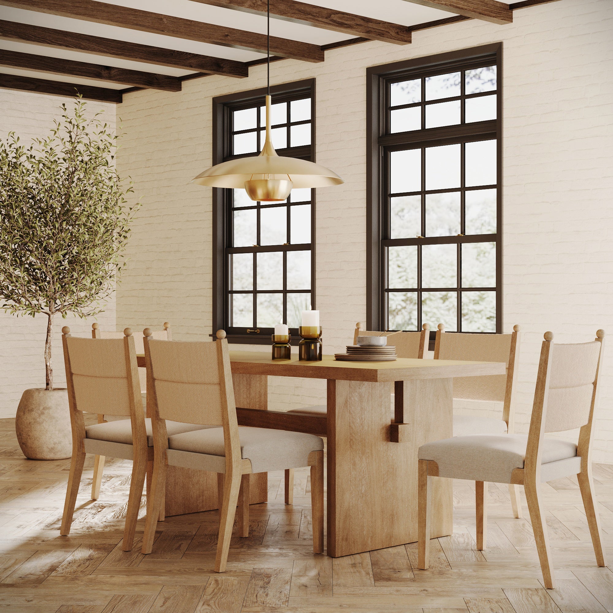 A cozy dining room with a wooden table, six upholstered chairs, a modern gold pendant light, candles, stacked books, a large potted plant, and two tall windows letting in natural light.