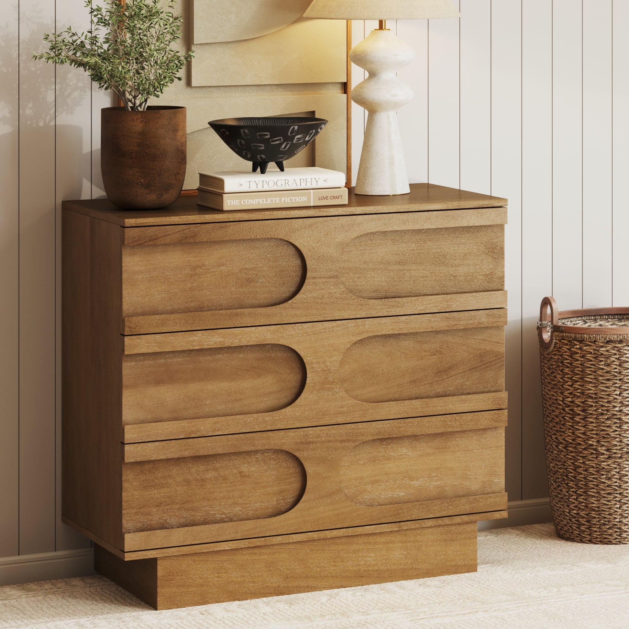 A wooden dresser with unique curved drawer details, topped with a potted plant, stacked books, a black bowl, and a white lamp. A woven basket sits on the floor beside it against a paneled wall.