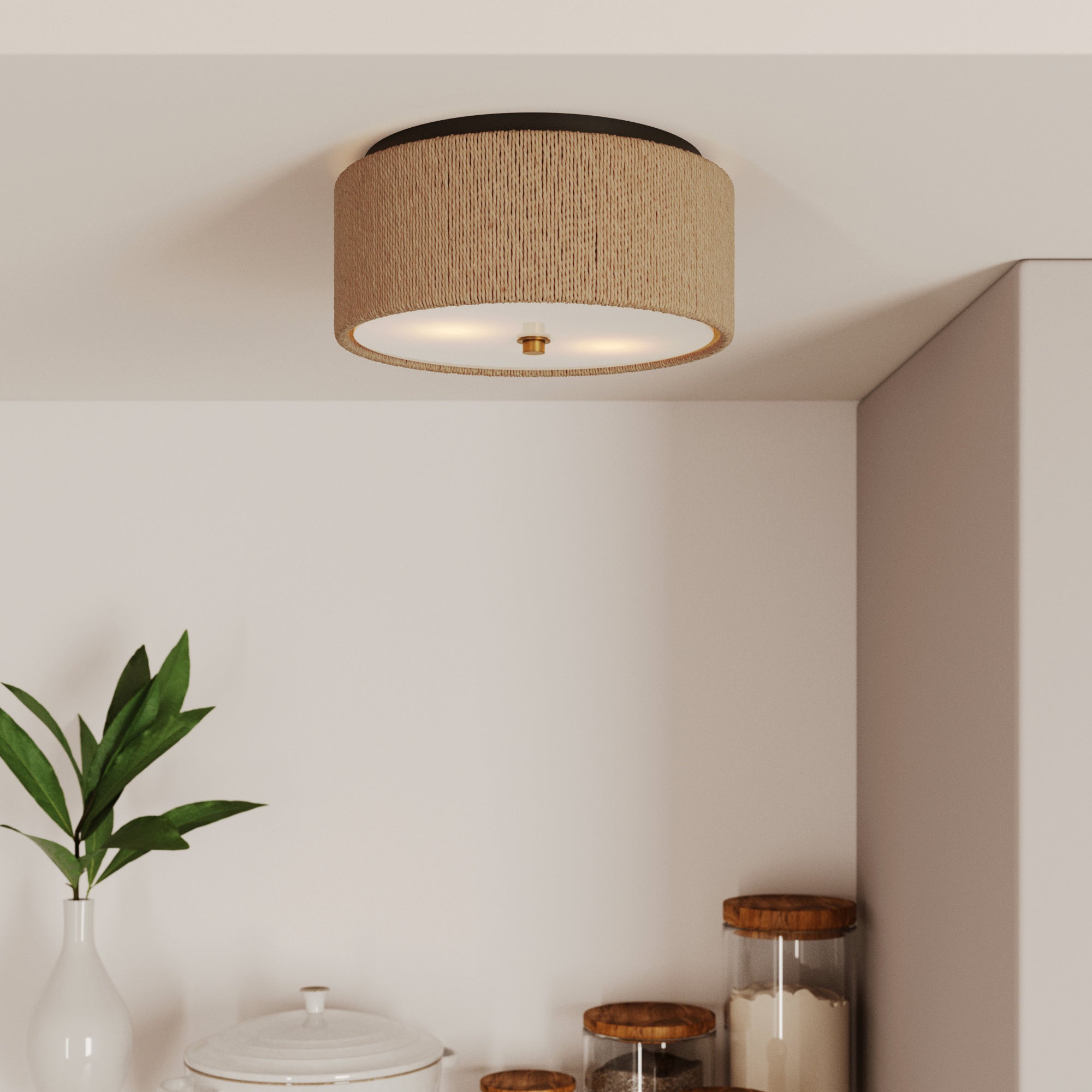 A Nathan James Seagrass Shade Flush Mount Ceiling Light with a round shape is mounted on a white kitchen ceiling above a counter holding glass jars with wooden lids, a white ceramic container, and a green leafy plant.