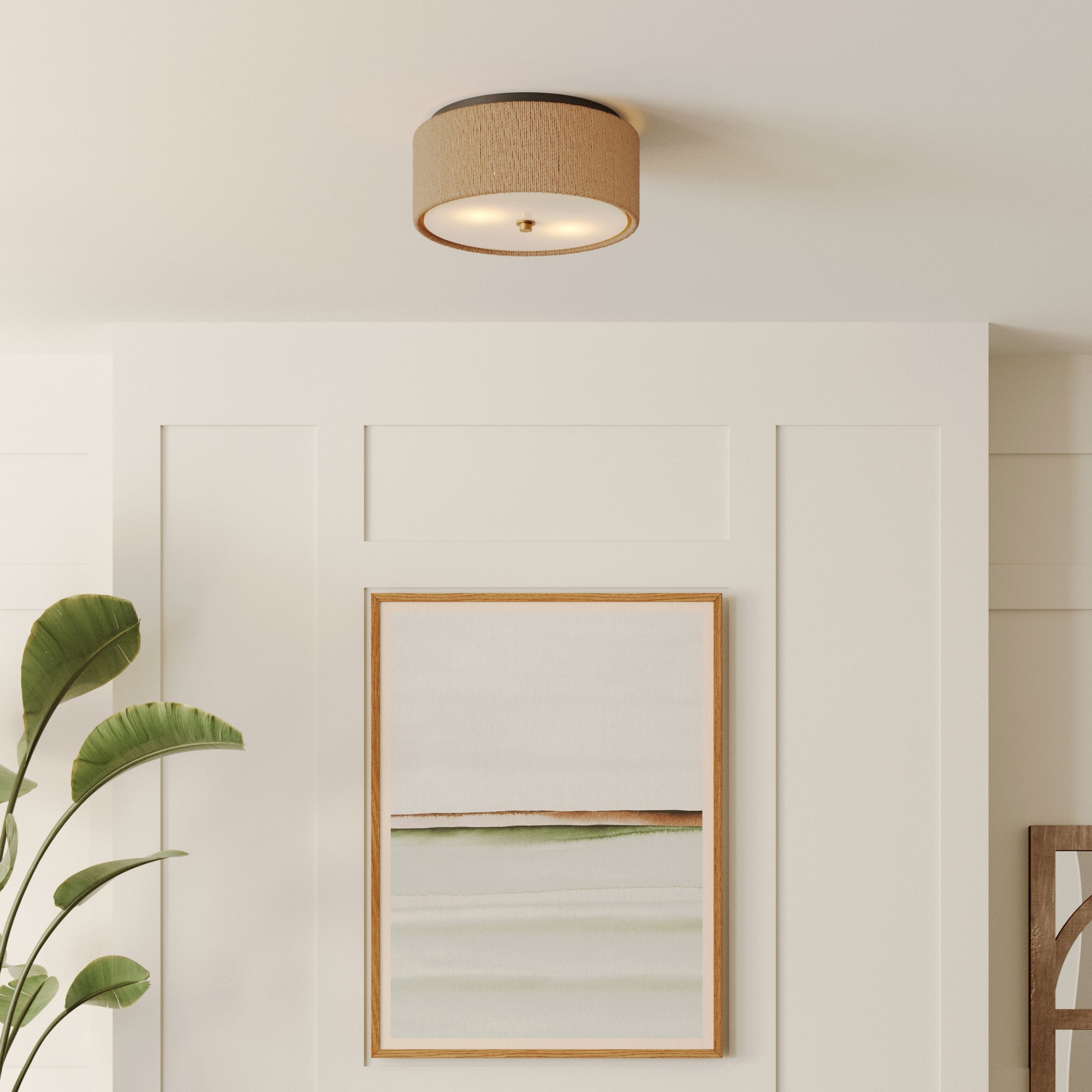 A modern, beige drum ceiling light fixture is mounted on a white paneled ceiling above a framed abstract painting with green and beige tones. A green leafy plant is visible on the left side.