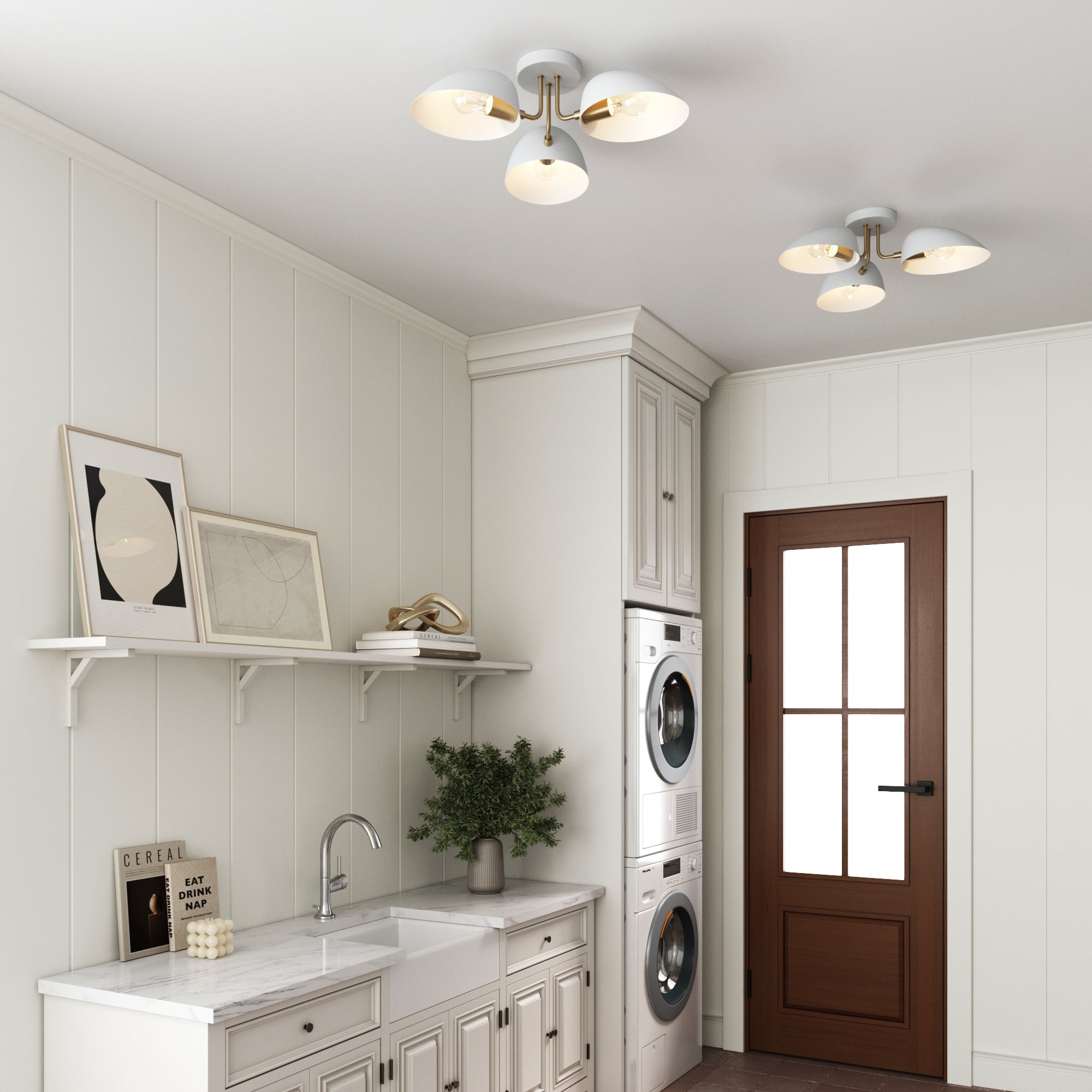 A bright, modern laundry room with white cabinets, wall art, plants, stacked washer and dryer, a sink, and a Nathan James Metal 3-Arm Flush Mount Ceiling Light (Set of 2). A wooden door with glass panels lets in natural light.