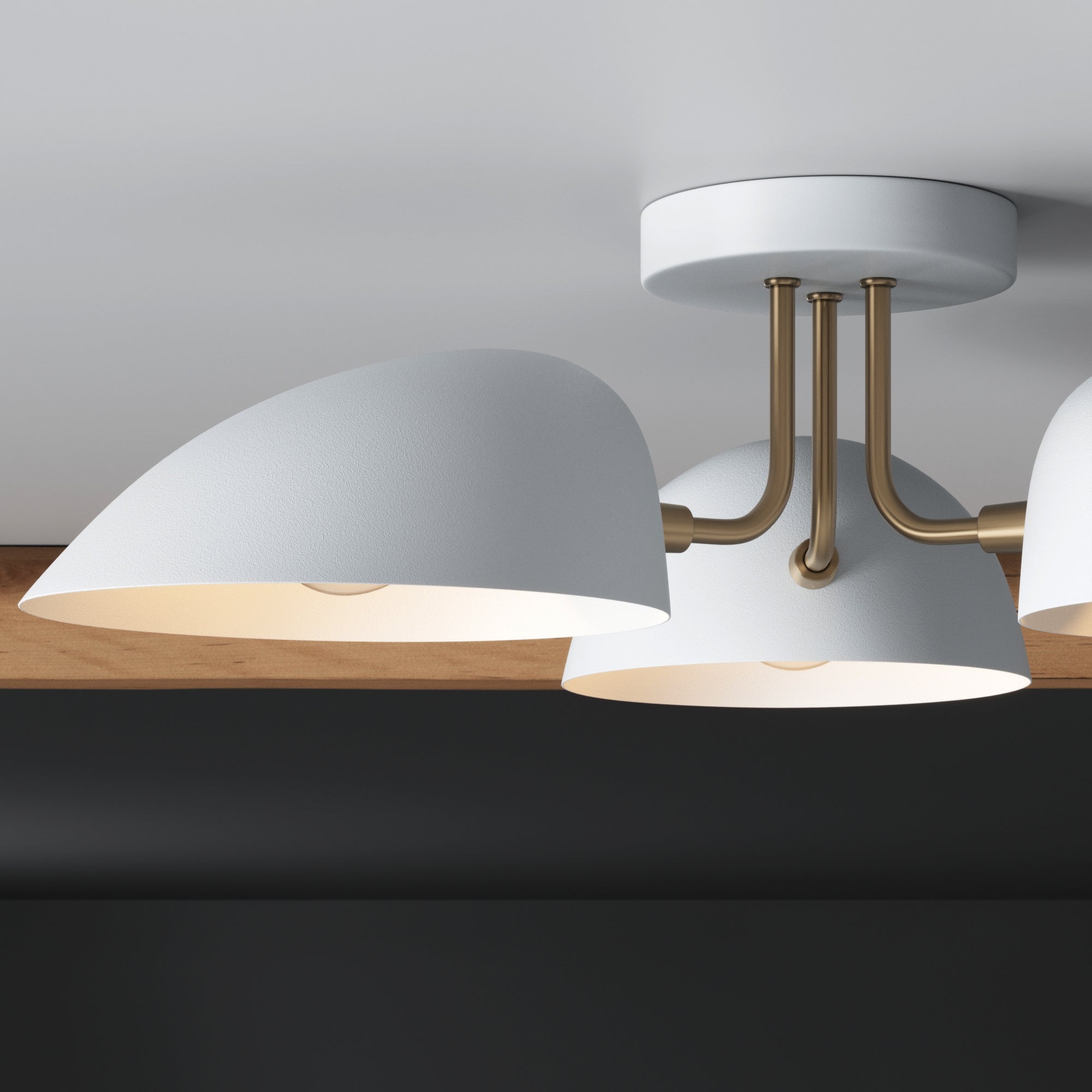 Close-up of a modern ceiling light fixture with three white, dome-shaped shades and brass arms, mounted on a white ceiling above a dark-colored wall.