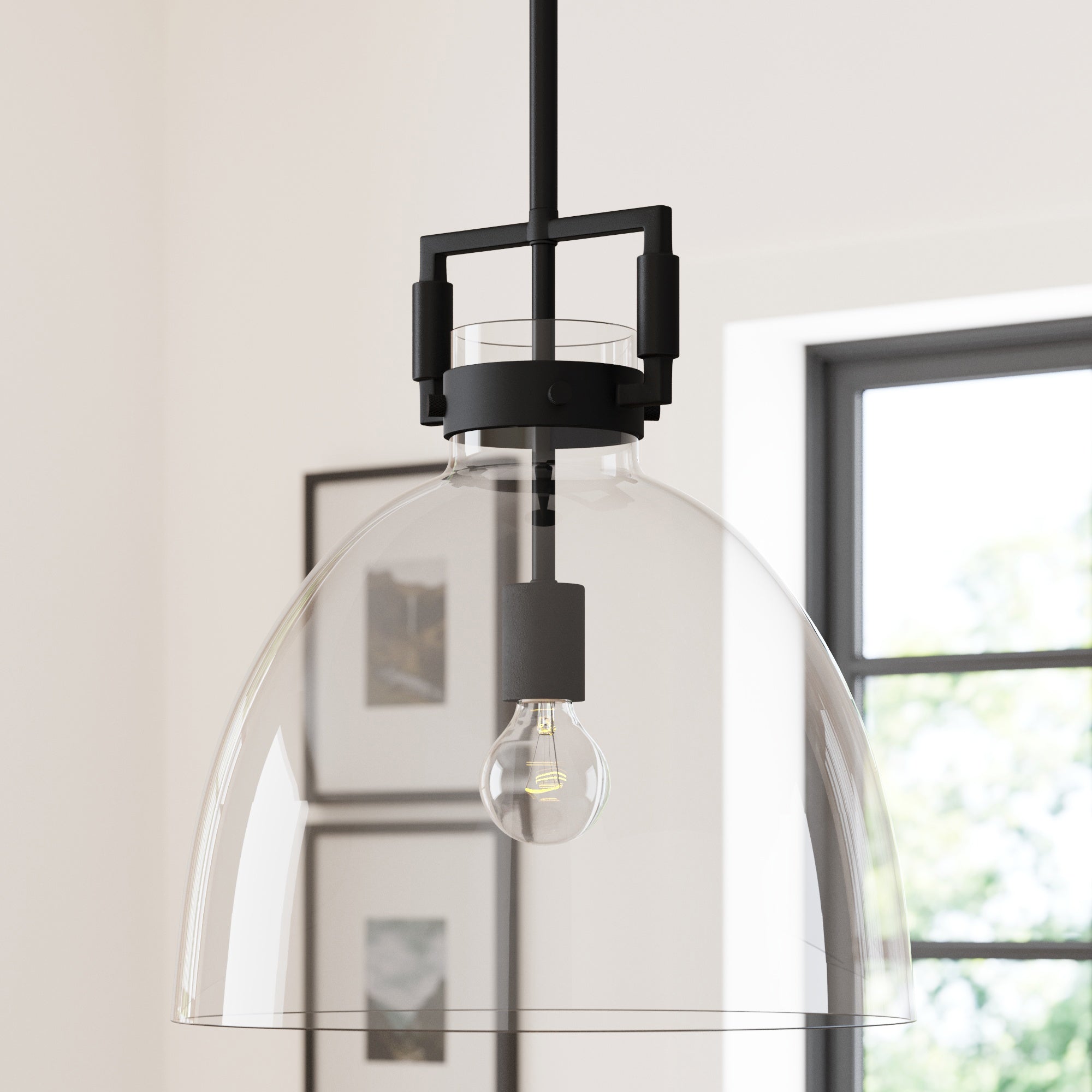A close-up of a modern pendant light with a clear glass dome shade and exposed light bulb, hanging from a black rod in a bright room with framed wall art and a window in the background.