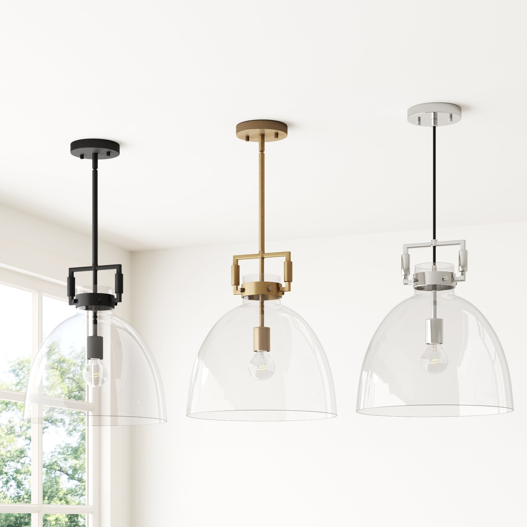 Three modern pendant lights with clear glass dome shades hang from the ceiling in a row, each with a different colored fixture: black, gold, and silver. A window with green trees outside is visible in the background.