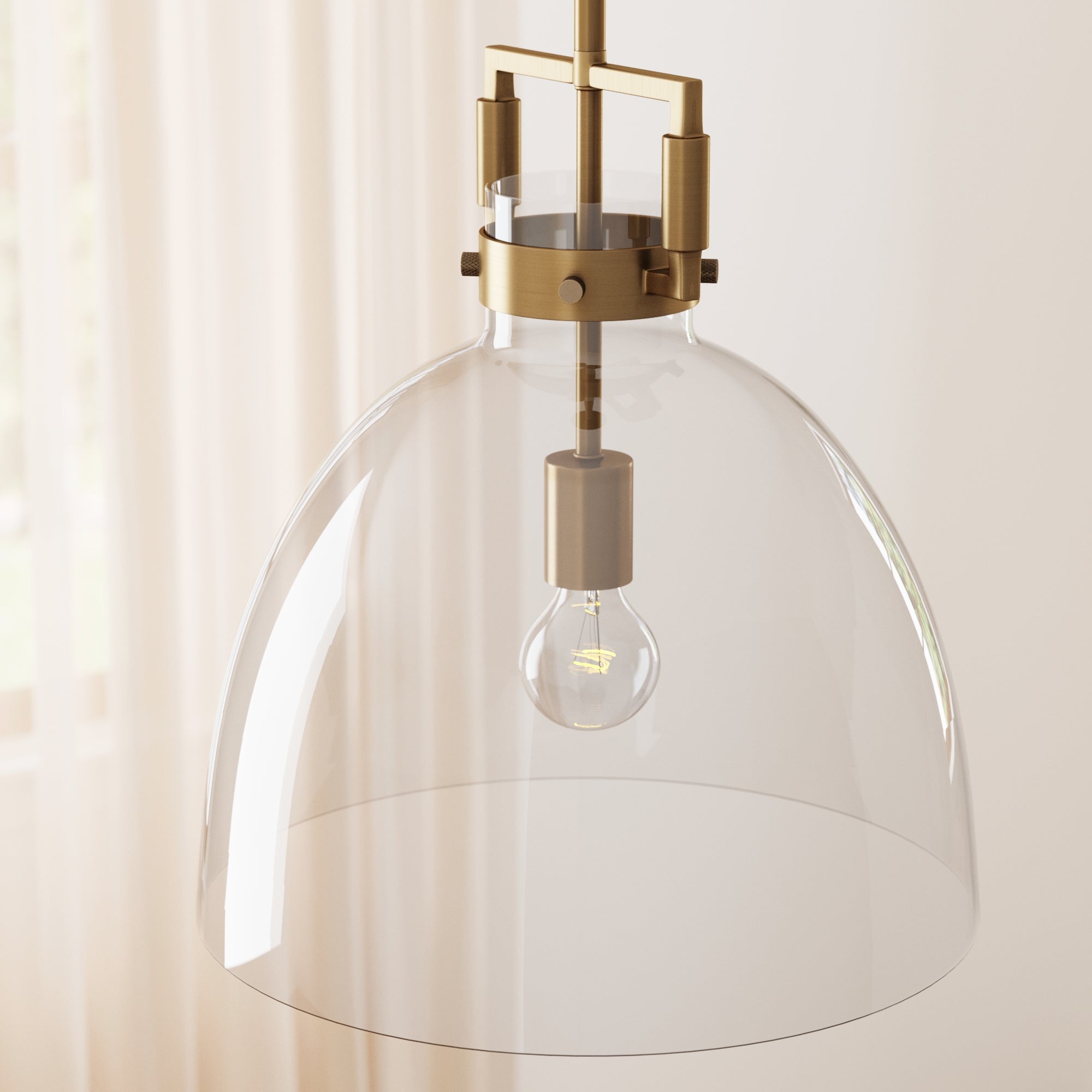 A modern pendant light with a clear glass dome shade, brass hardware, and a visible light bulb, hanging in a softly lit room with sheer curtains in the background.