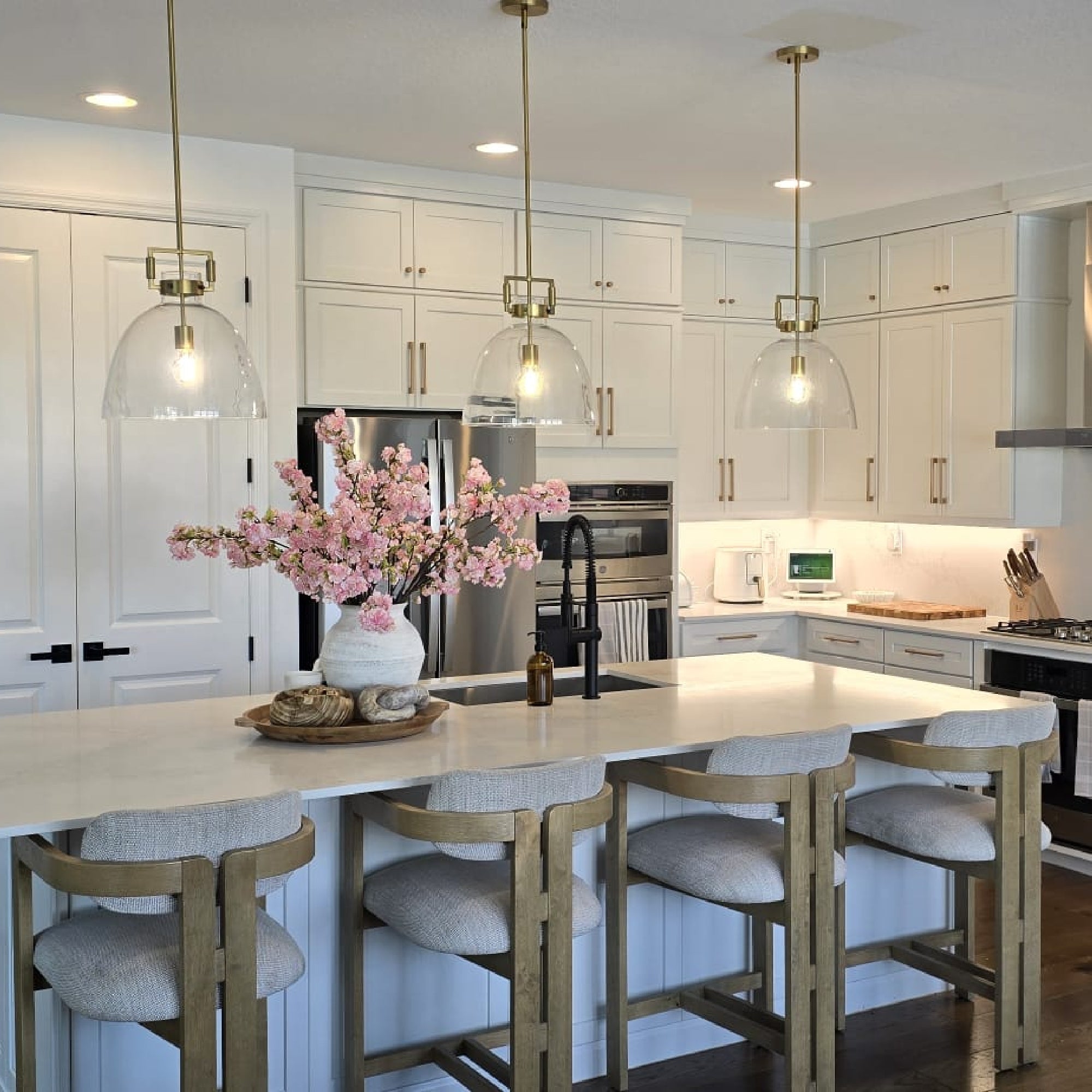 A modern kitchen with white cabinets, a large island with four upholstered stools, pendant lights, stainless steel appliances, and a vase of pink flowers as a centerpiece on the counter.