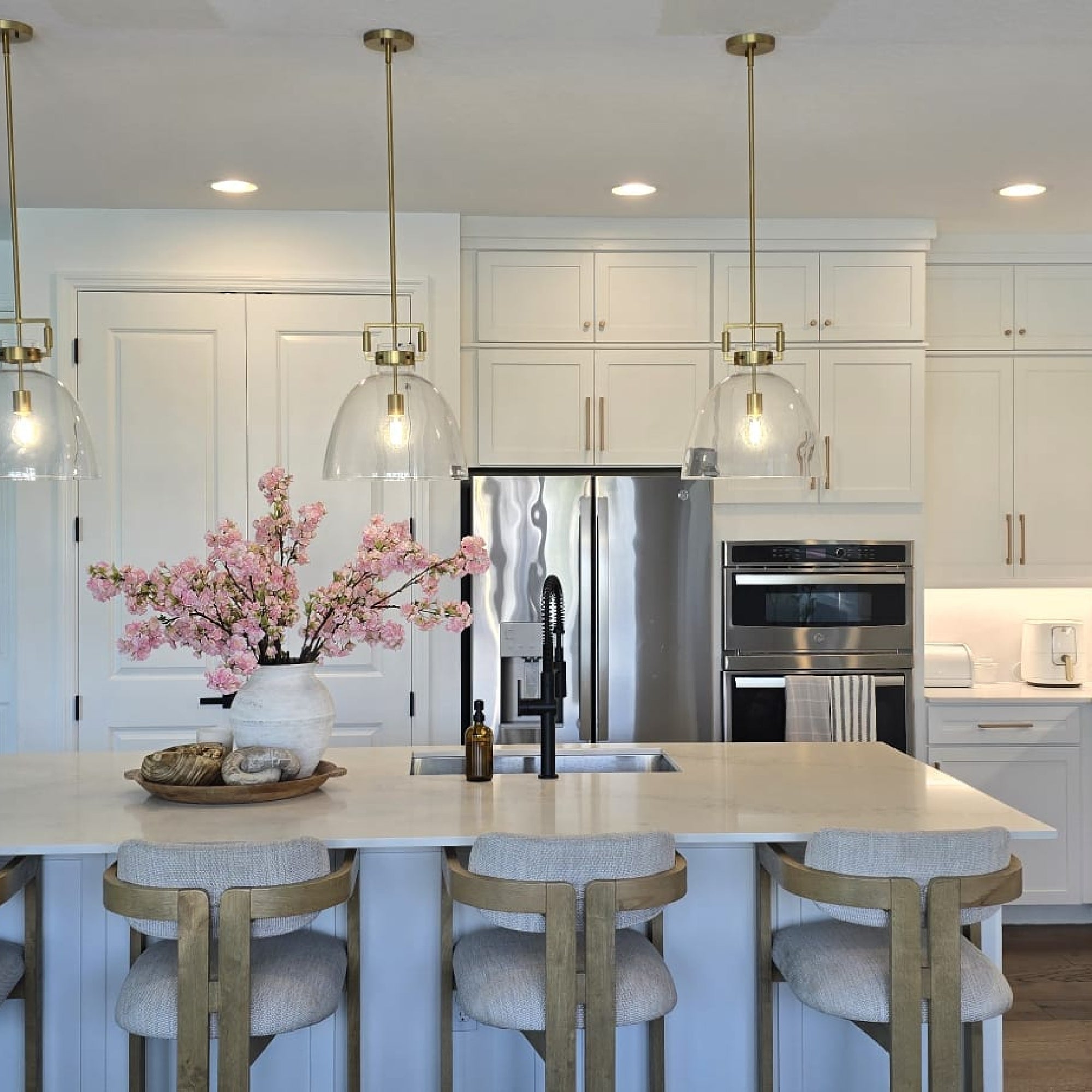 A modern kitchen with white cabinets, stainless steel appliances, and a central island with four cushioned stools. Three pendant lights hang above, and a vase of pink flowers decorates the counter.