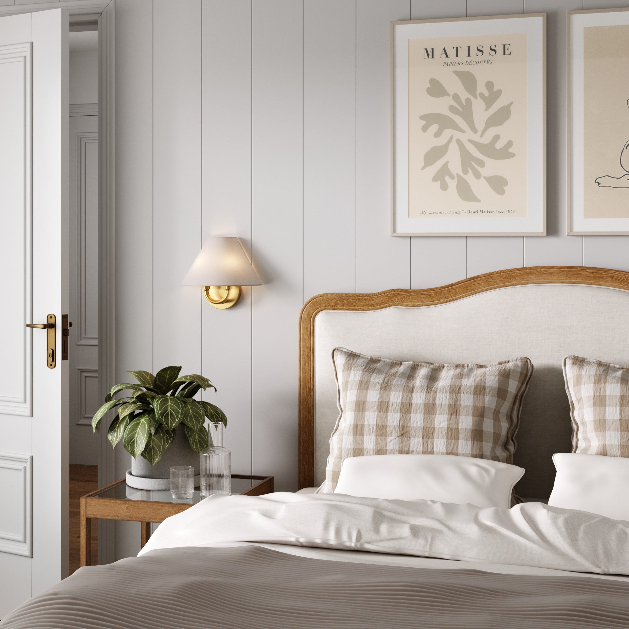 A cozy bedroom features a wooden headboard, checkered pillows, and white bedding. A plant sits on the bedside table, while the Nathan James Modern Farmhouse Wall Light with Linen Shade softly illuminates art prints above the bed on a gray paneled wall.