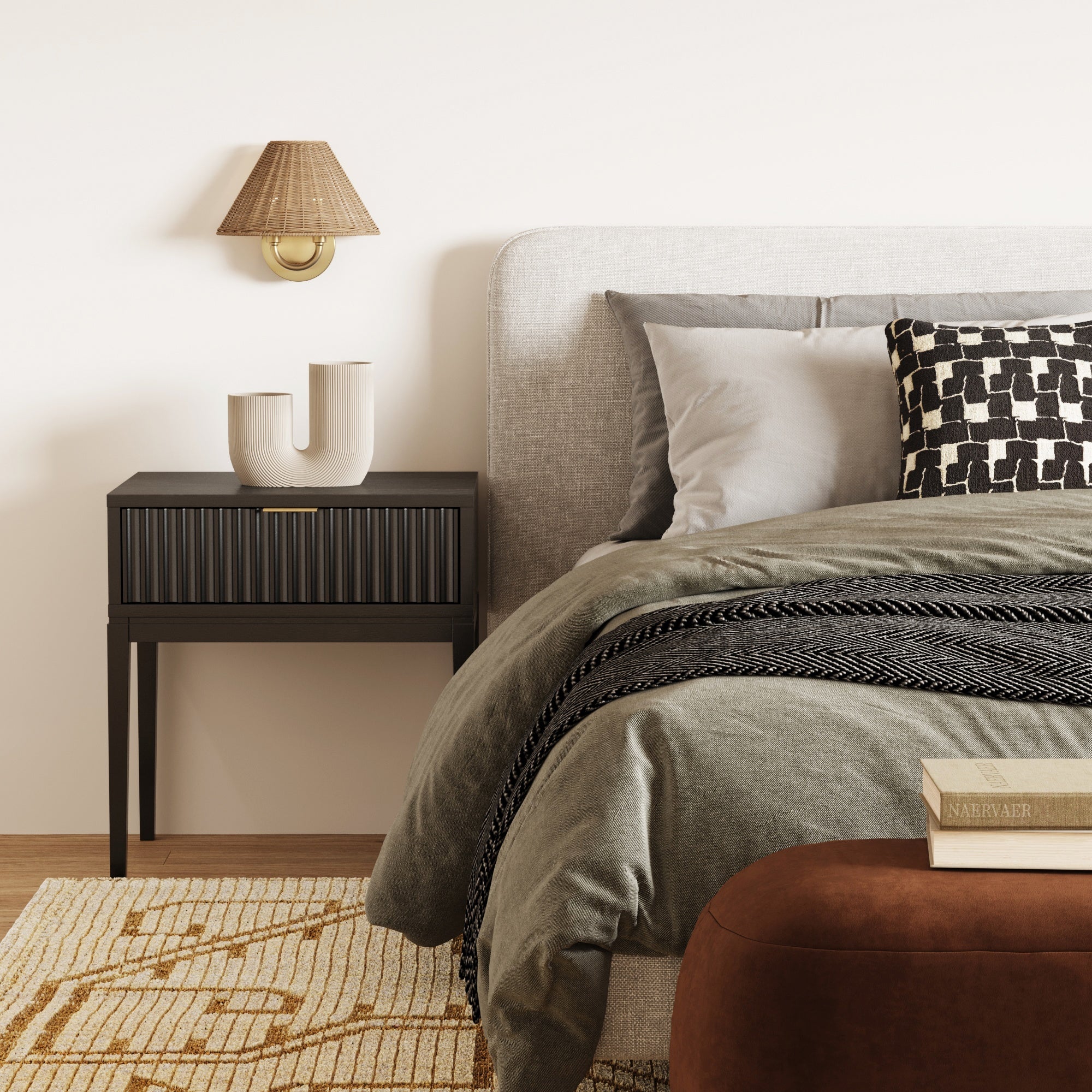 A modern bedroom with a gray upholstered bed, black-and-white patterned pillow, greenish-gray bedding, black nightstand with a ceramic vase, Nathan James Modern Farmhouse Wall Light with Rattan Shade, and a brown bench on a beige geometric rug.