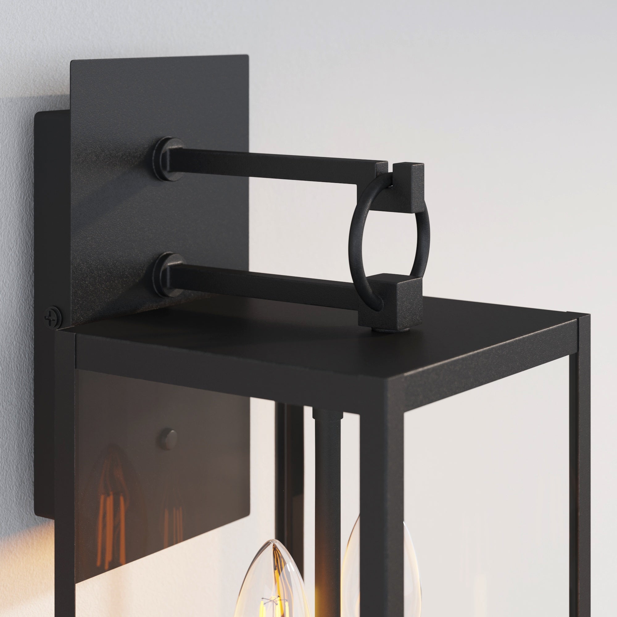 A close-up of a modern black metal outdoor wall lantern with a rectangular backplate, clear glass panels, and geometric detailing, mounted on a light gray wall.