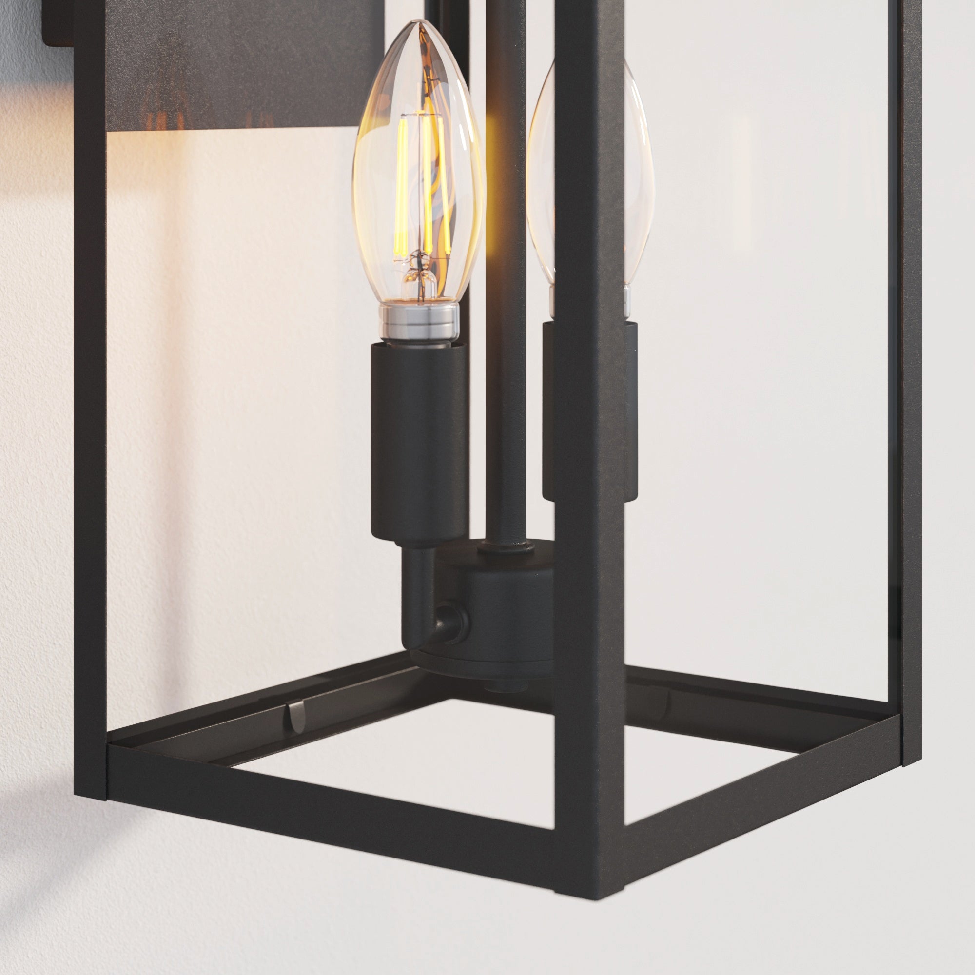 Close-up of a modern black metal wall sconce with a clear glass enclosure, showcasing an exposed candle-shaped LED bulb mounted on a black fixture against a white wall.