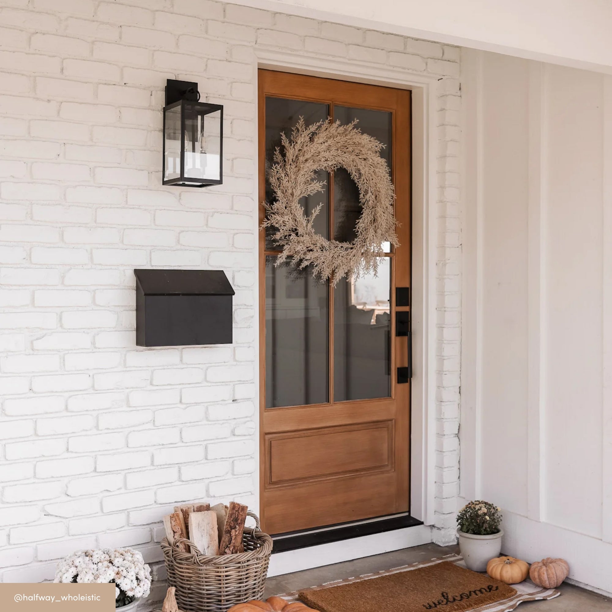 A wooden front door with a glass panel is decorated with a wreath. The white brick exterior features a black lantern and mailbox. A doormat, basket of firewood, and pumpkins are on the porch, creating a cozy autumn look.