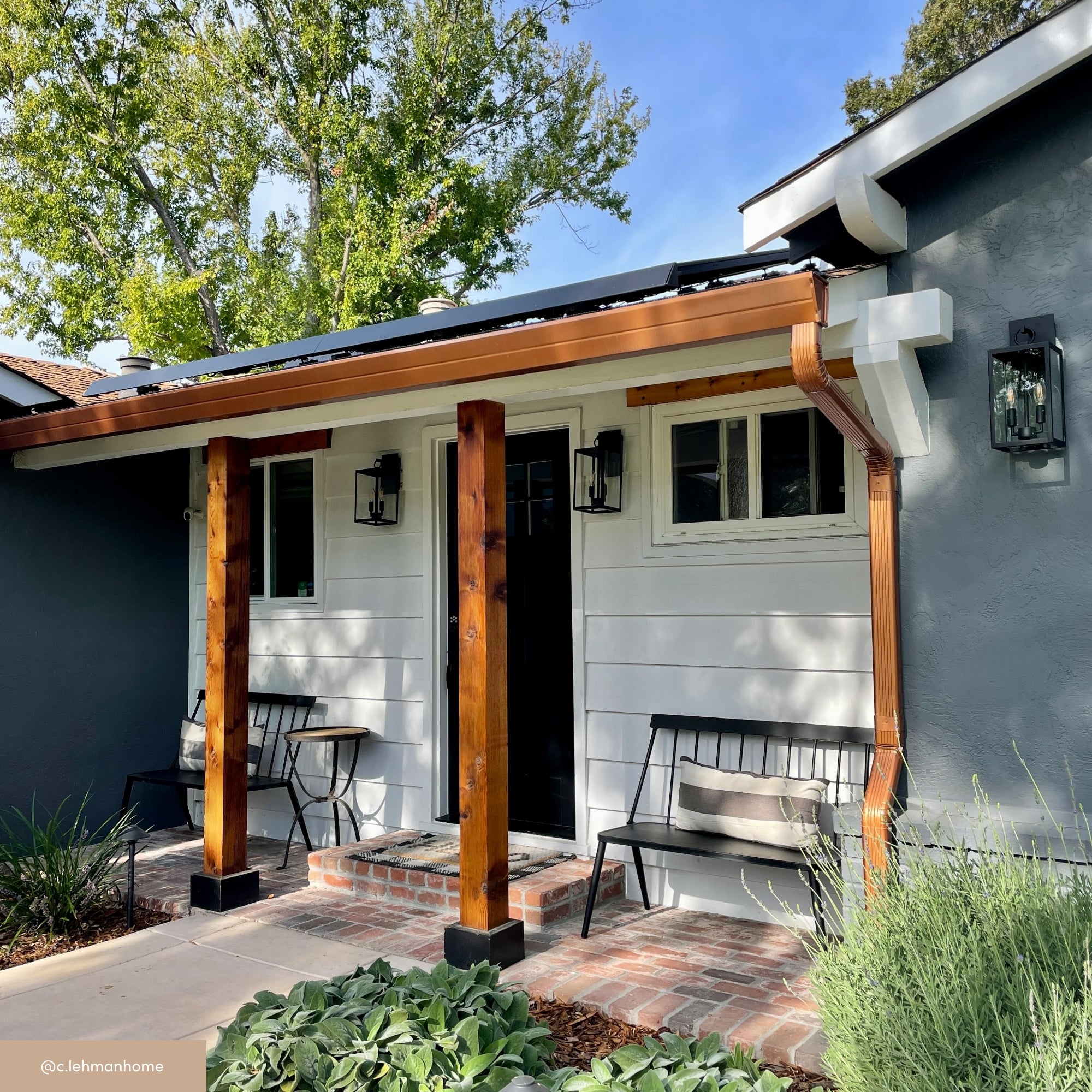 A modern porch with wooden beams, black benches, and outdoor lanterns decorates a gray house with white trim. Brick steps lead to a black front door, surrounded by greenery and trees.
