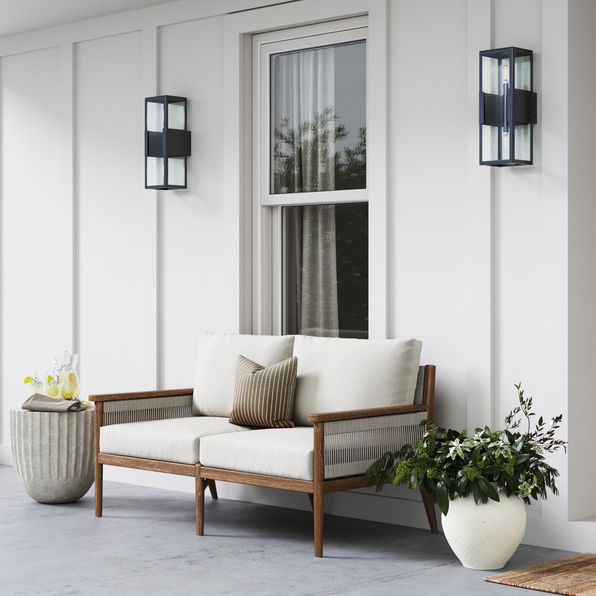 A modern patio with a wooden loveseat featuring cream cushions and a striped pillow, a round side table with drinks, two black wall lanterns, and a white plant pot with green foliage in front of a window.
