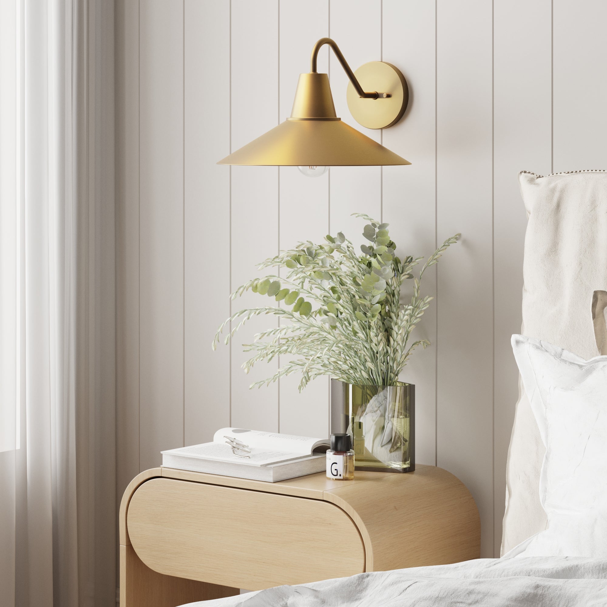 A modern bedroom features a light wood nightstand with a glass vase of greenery, white book, and small bottle. Above is the Nathan James Mid-Century Modern Metal Vintaged Brass Wall Sconce; part of a white bed is seen on the right.