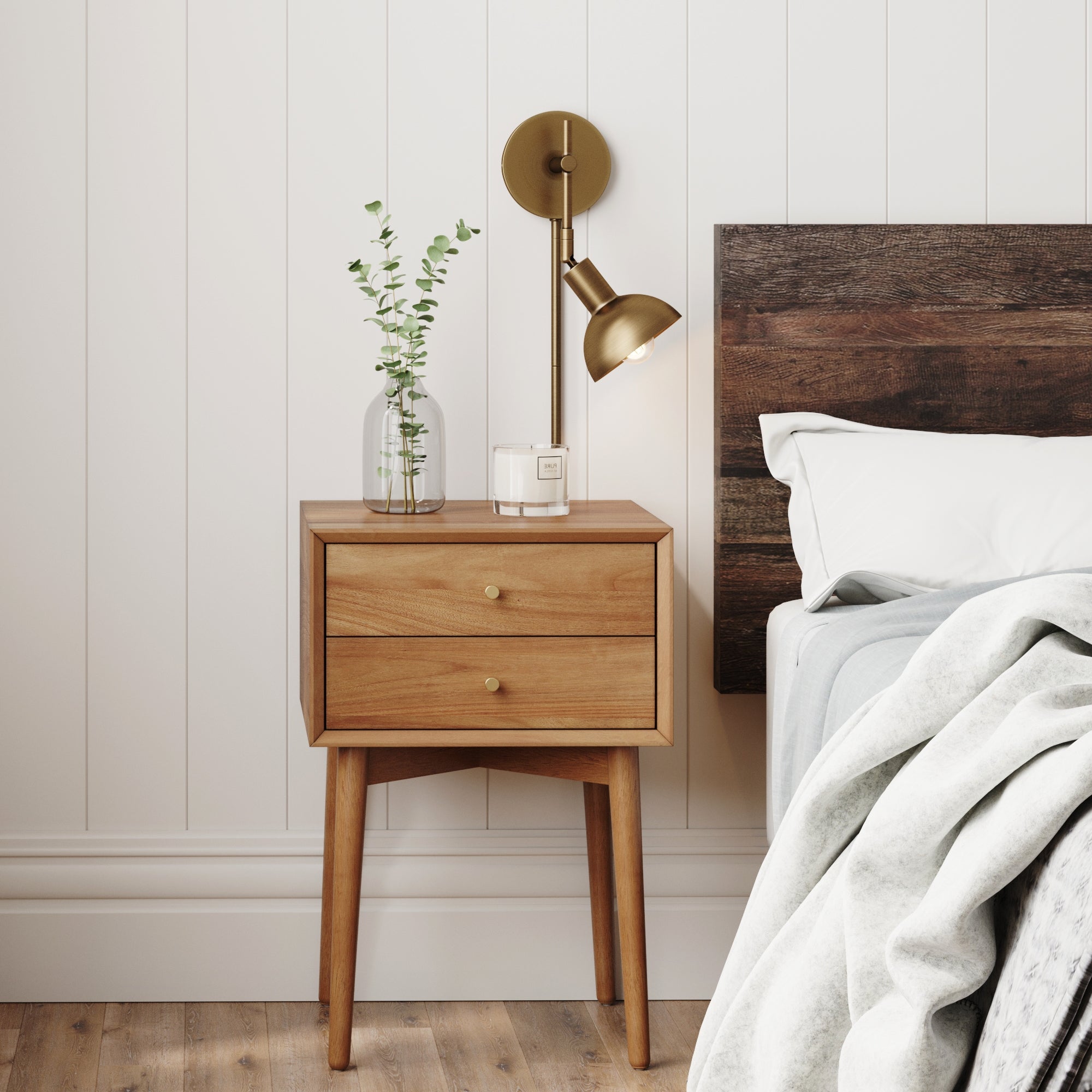 A wooden nightstand with two drawers holds a glass vase with greenery and a small box. A brass wall sconce is mounted above, beside a wooden bed with white bedding and a gray blanket on a wooden floor.