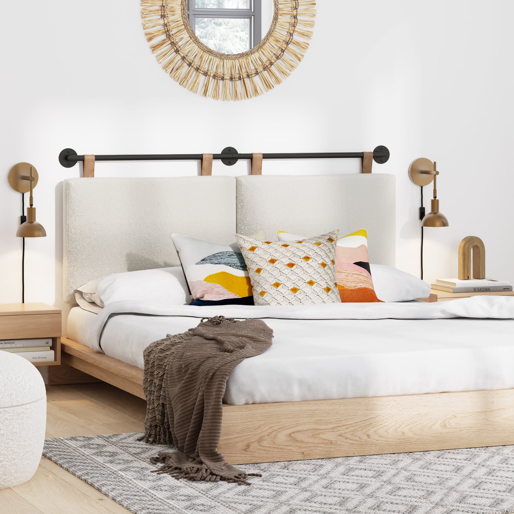 A modern bedroom with a light wood bed, white upholstered headboard, colorful throw pillows, a brown blanket, bedside tables with lamps, a patterned rug, and a round decorative mirror on the wall.