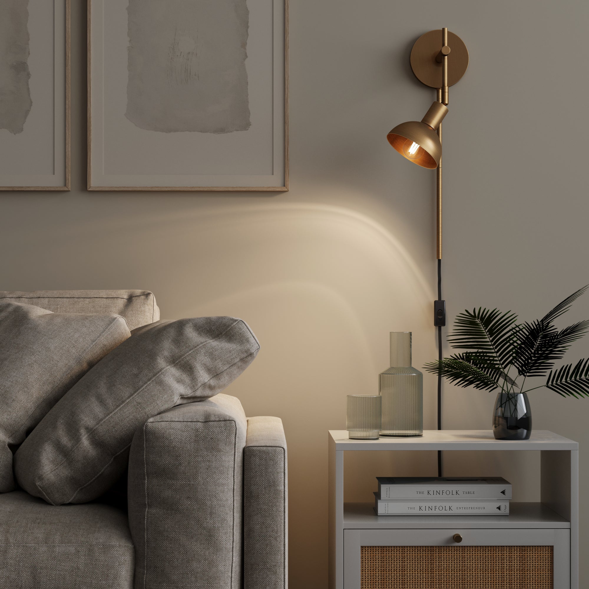 A modern living room corner with a gray sofa, a white side table holding books, two glass vases, a green plant, and a gold wall-mounted lamp illuminating the beige wall with framed art above the sofa.