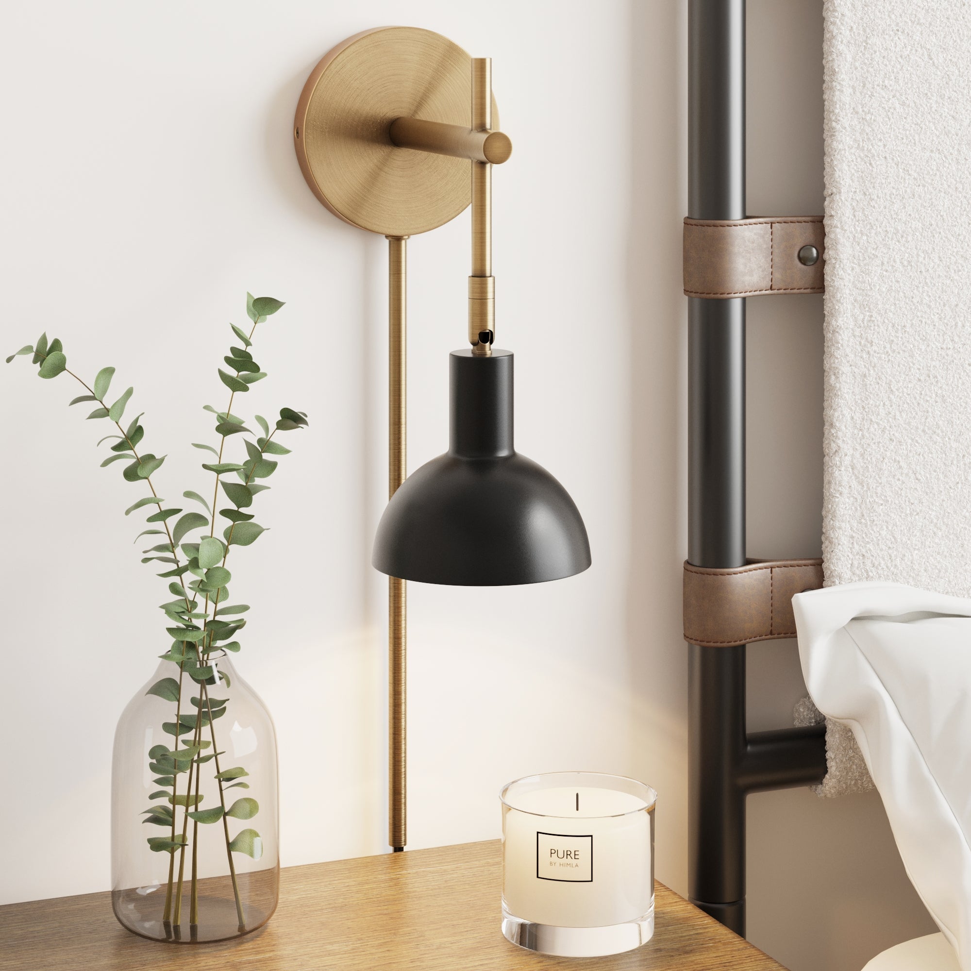 A modern bedside features a glass vase with eucalyptus, a lit PURE candle, and the Nathan James Plug-In Adjustable Metal Wall Light in Black Brass above. Part of a white-bedded bed and a towel on a leather-strap ladder are visible.