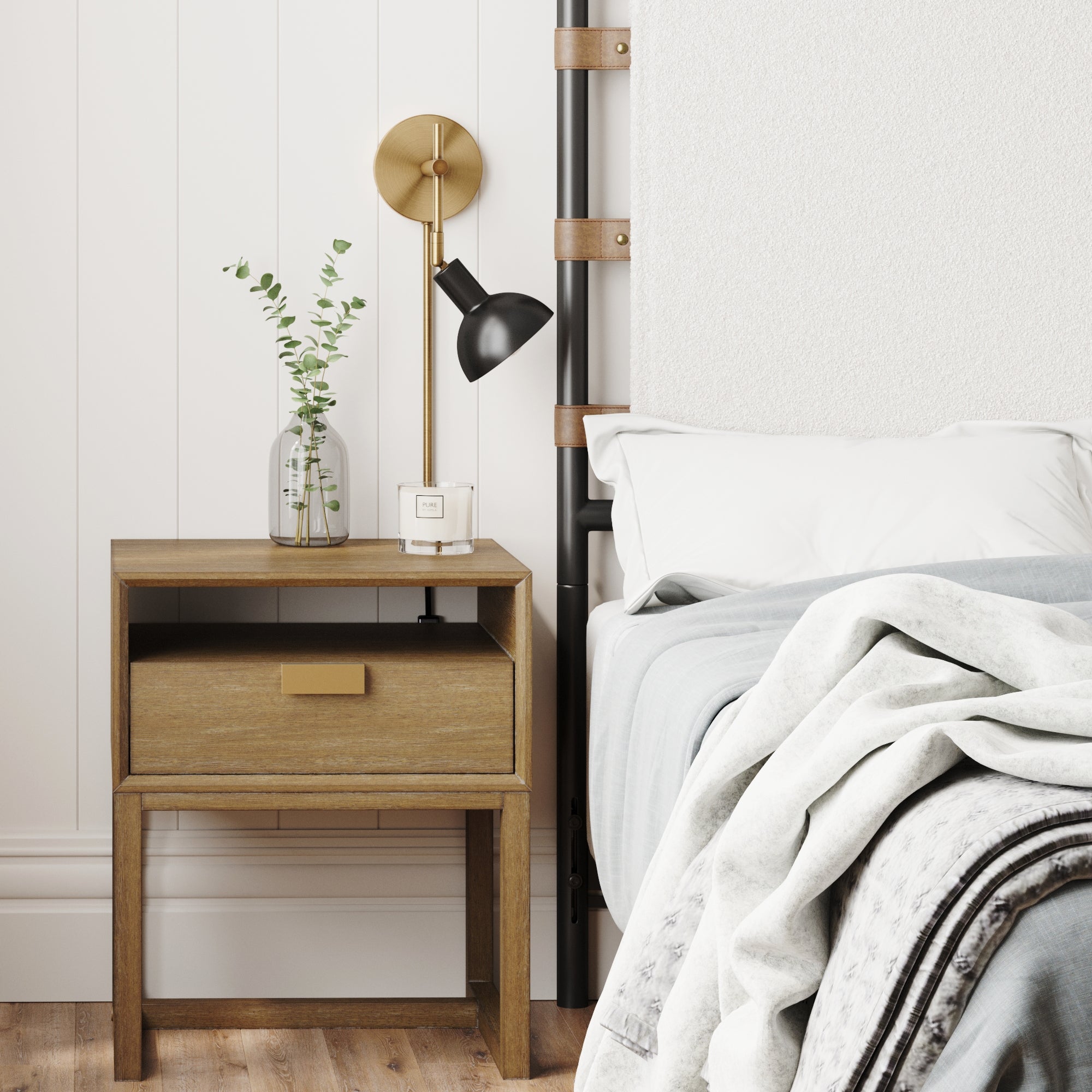 A cozy bedroom with a wooden nightstand holding a glass vase with green leaves and a small white clock, beside a neatly made bed with white and gray bedding. A gold and black wall lamp is above the nightstand.