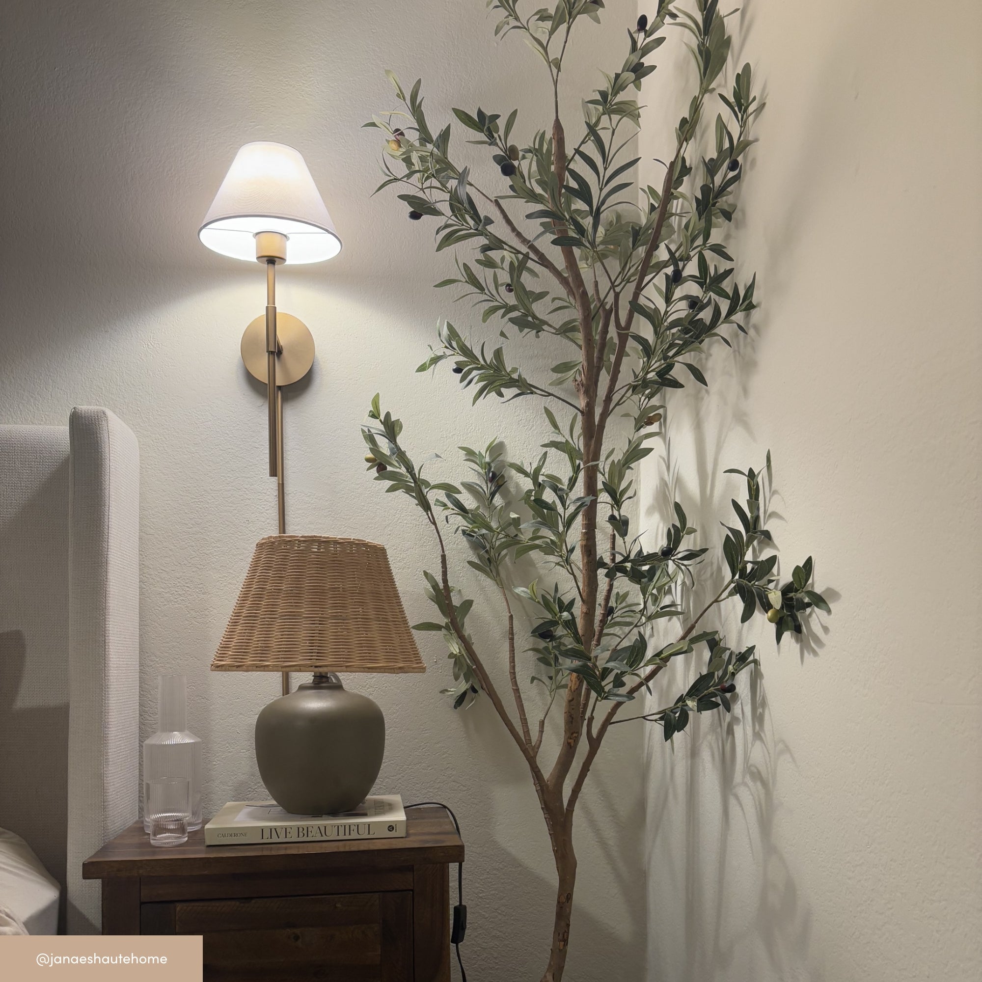 A cozy bedroom corner with a wall-mounted lamp, a leafy potted tree, a bedside table holding a wicker-shaded lamp, a glass of water, and books, including one titled LIVE BEAUTIFULLY.