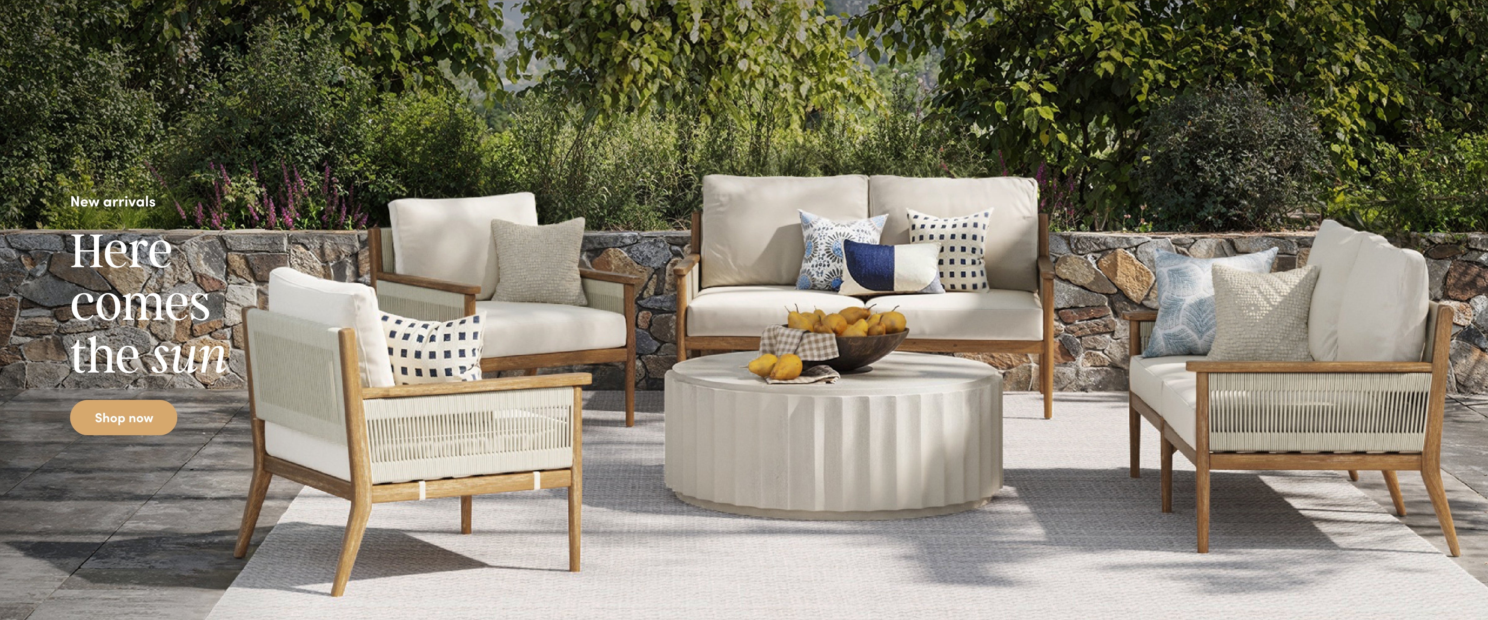 Outdoor patio set with four cushioned armchairs, a cushioned loveseat, and a round coffee table topped with a fruit bowl, all set on a rug with greenery and a stone wall in the background. Text: “Here comes the sun.”.