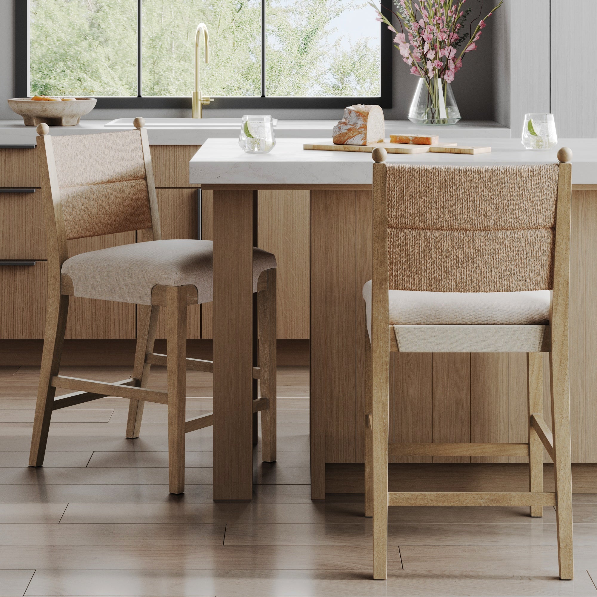 A modern kitchen features Nathan James Seagrass & Wood Counter Height Bar Stools in light brown at a wood island with marble countertop, glass cups with lime, bread on a cutting board, a bowl, flowers in a vase, and a window showing greenery outside.