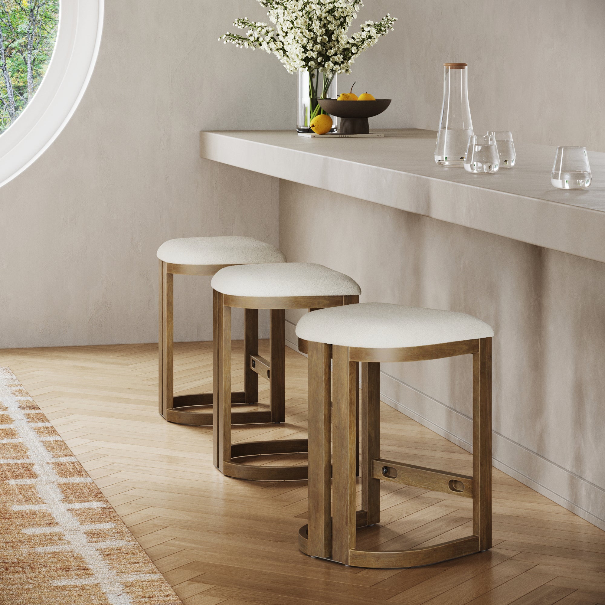 Three Nathan James Barrel Wooden Backless Bar Stools in light brown are lined up by a sleek, pale countertop topped with a vase of white flowers, lemons, and glassware. A round window serves as the scenic backdrop. Set includes 3 stools.