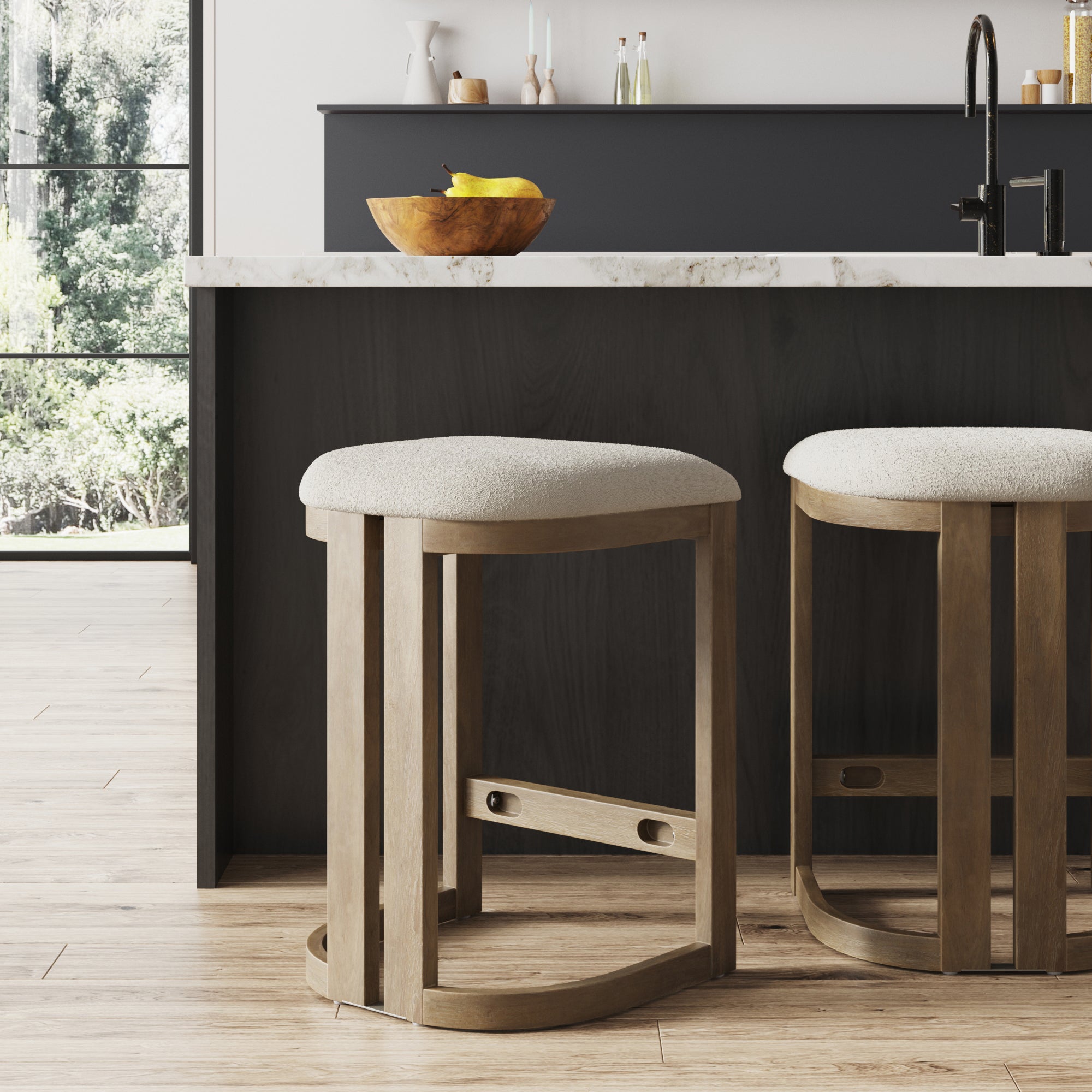 Two Nathan James Barrel Wooden Backless Bar Stools with upholstered seats are at a modern kitchen island with a marble countertop, bananas in a bowl, and a tap; sunlight streams through large windows onto wooden floors.