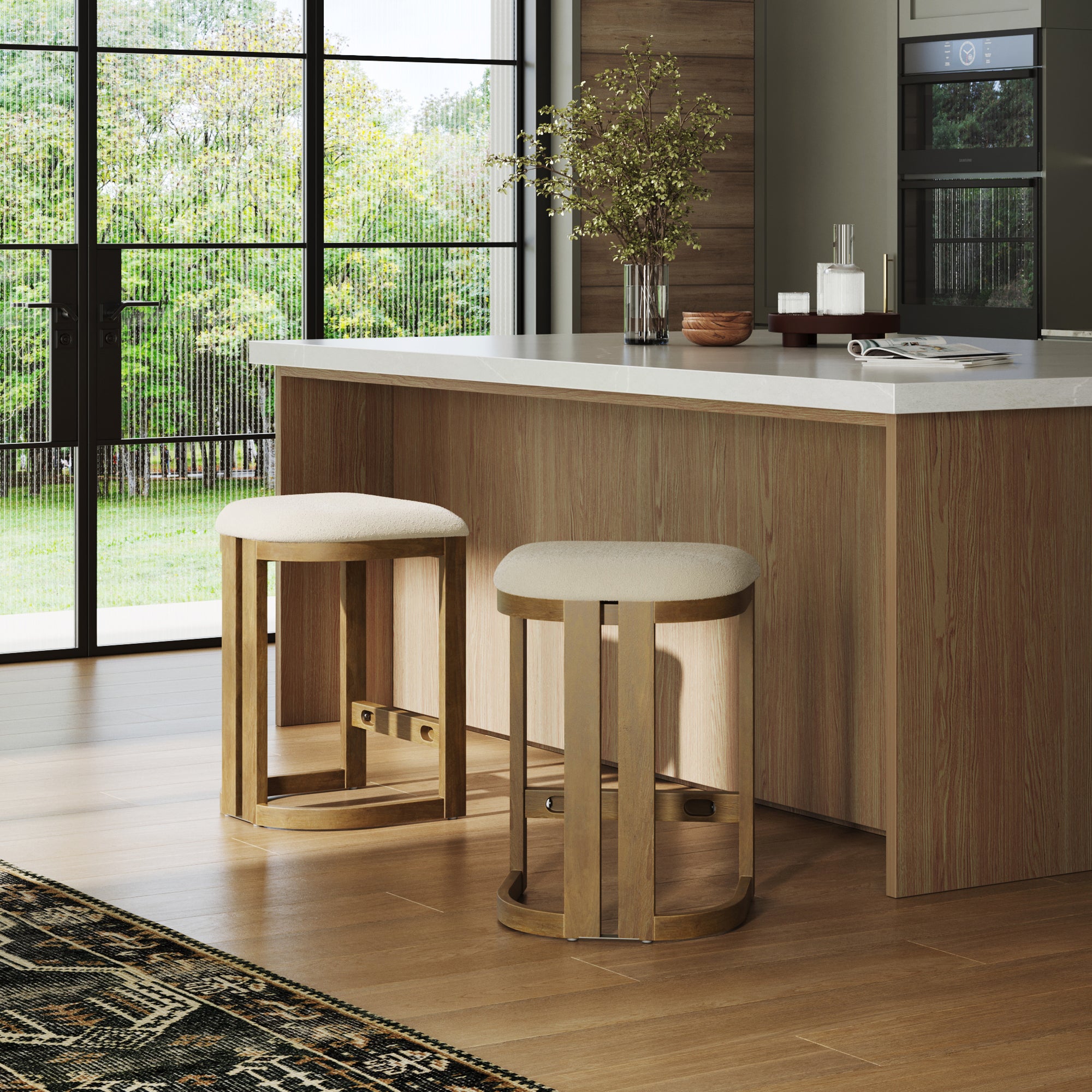 A modern kitchen features a solid wood island with a white countertop, two Nathan James Barrel Wooden Backless Bar Stools in light brown with upholstered seats, a patterned rug, decorative accents, and large windows overlooking the green yard.