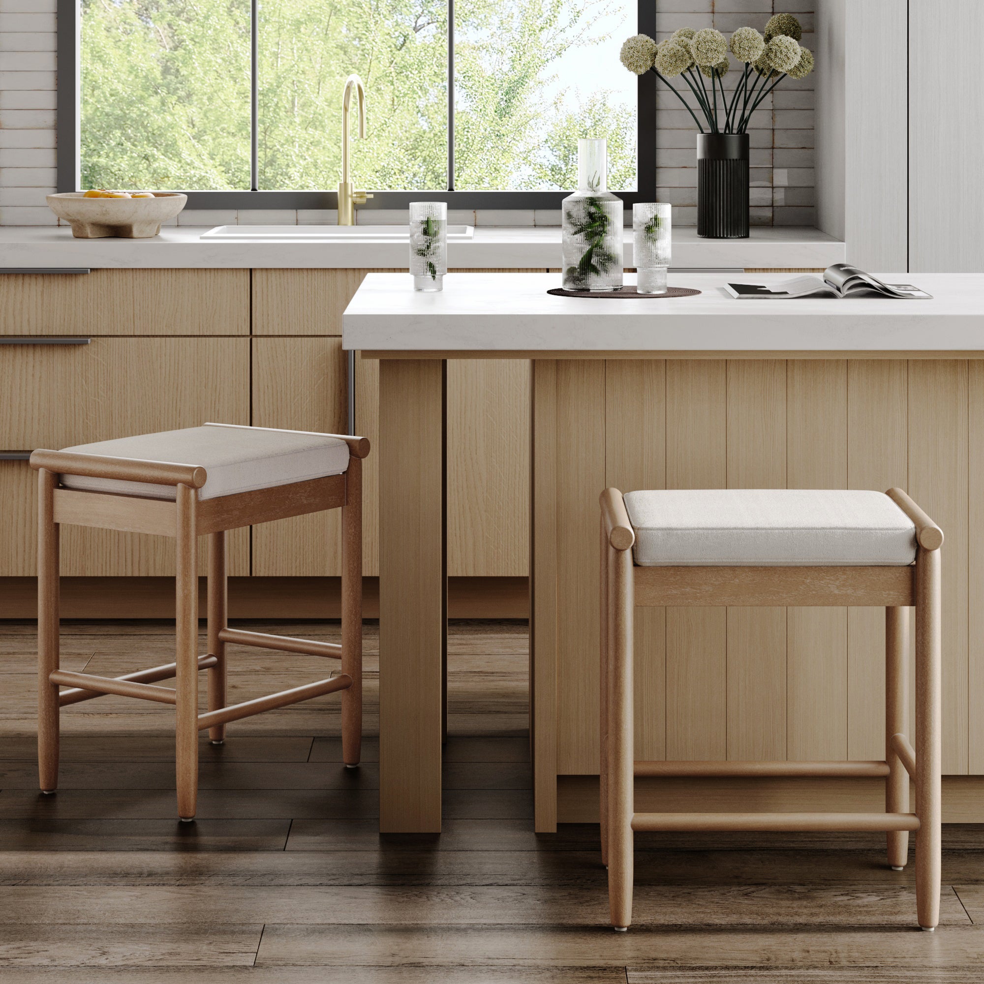 A modern kitchen showcases light wood cabinets, a white island with two Nathan James Backless Wooden Bar Stools with Boucle Upholstery in Light Brown, a large window overlooking greenery, and decorative items on the counter.