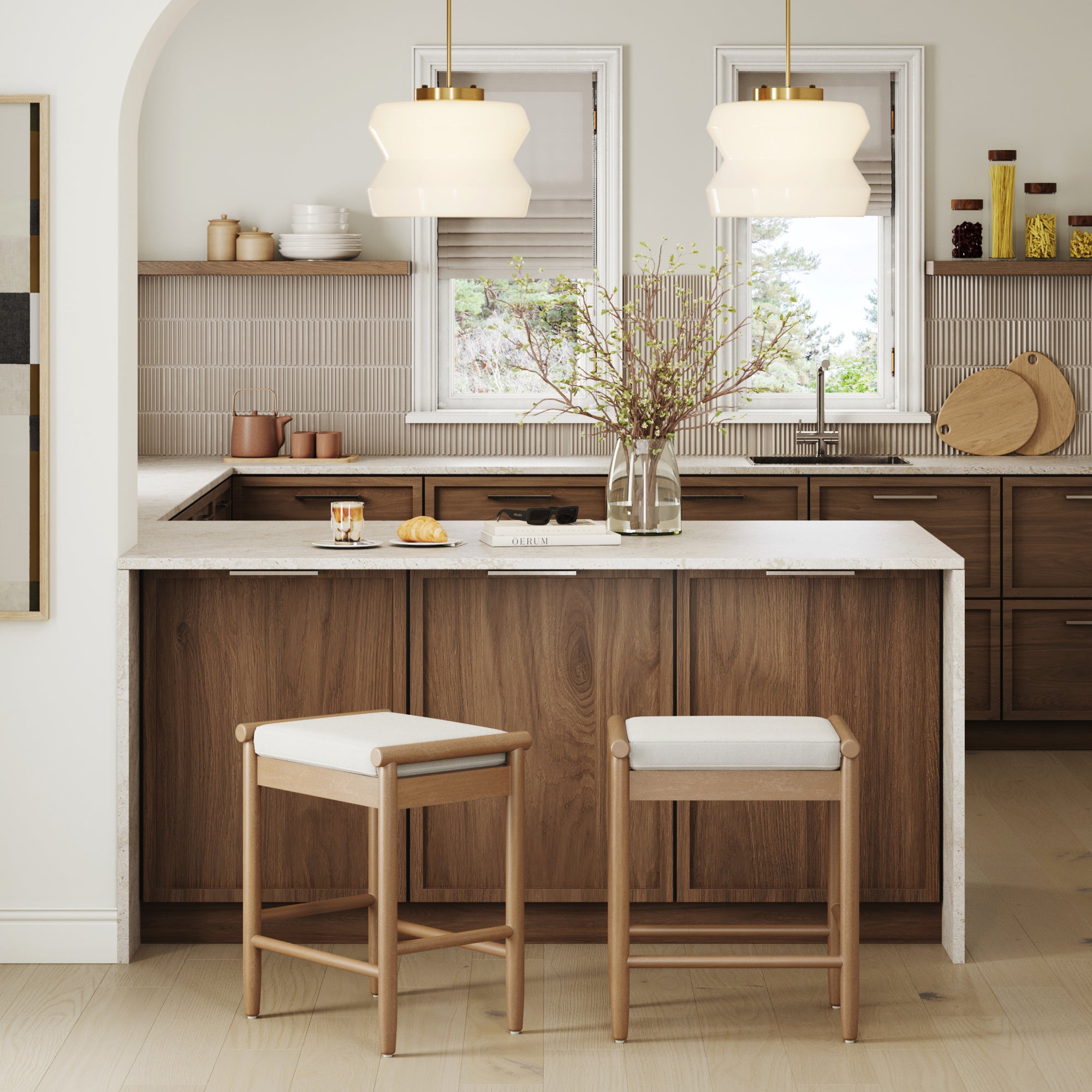 Modern kitchen with wooden cabinets, white countertops, and two Nathan James Backless Wooden Bar Stools with Boucle Upholstery in Light Brown. Pendant lights and a vase with branches accent the cozy, neutral space filled with natural light.