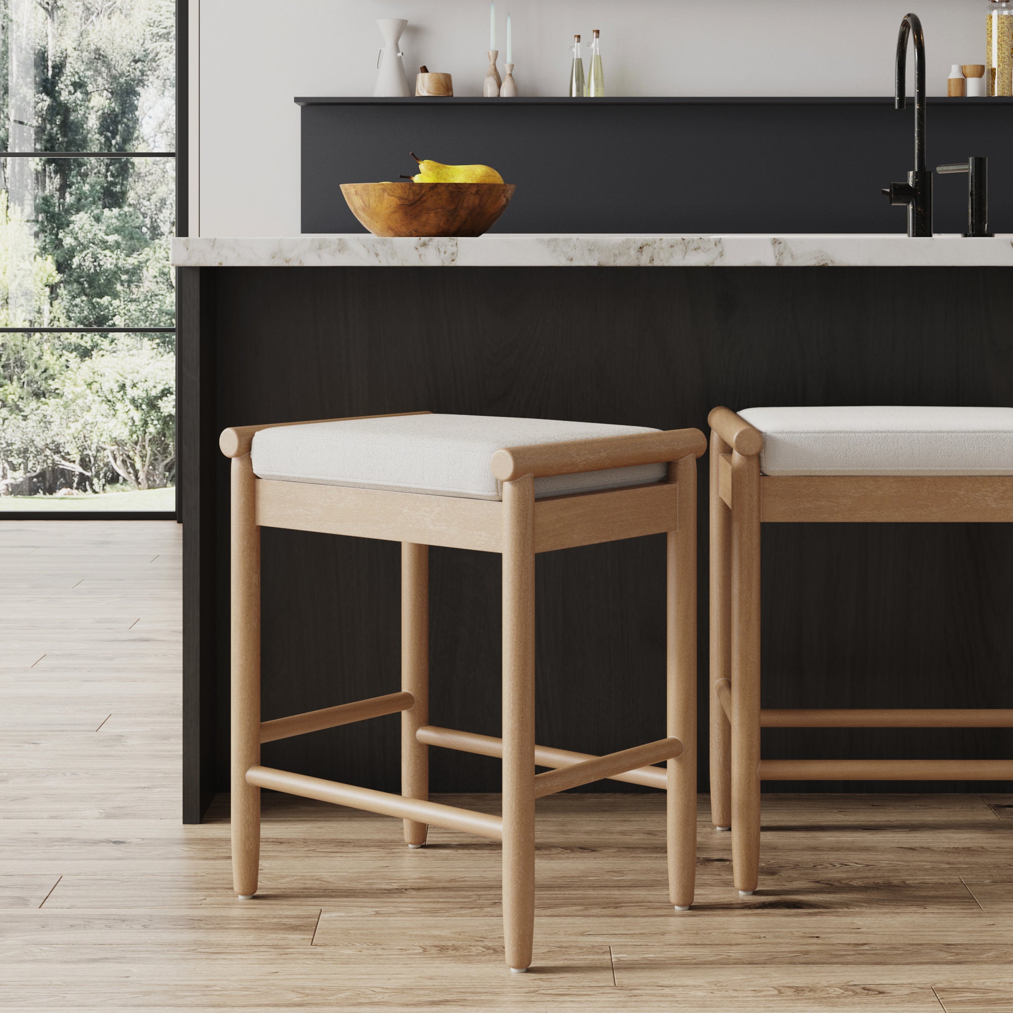 Two Nathan James Backless Wooden Bar Stools in light brown with boucle upholstery sit at a modern kitchen island with a dark base and marble countertop. Sunlight streams in, illuminating a wooden bowl of bananas on the counter.