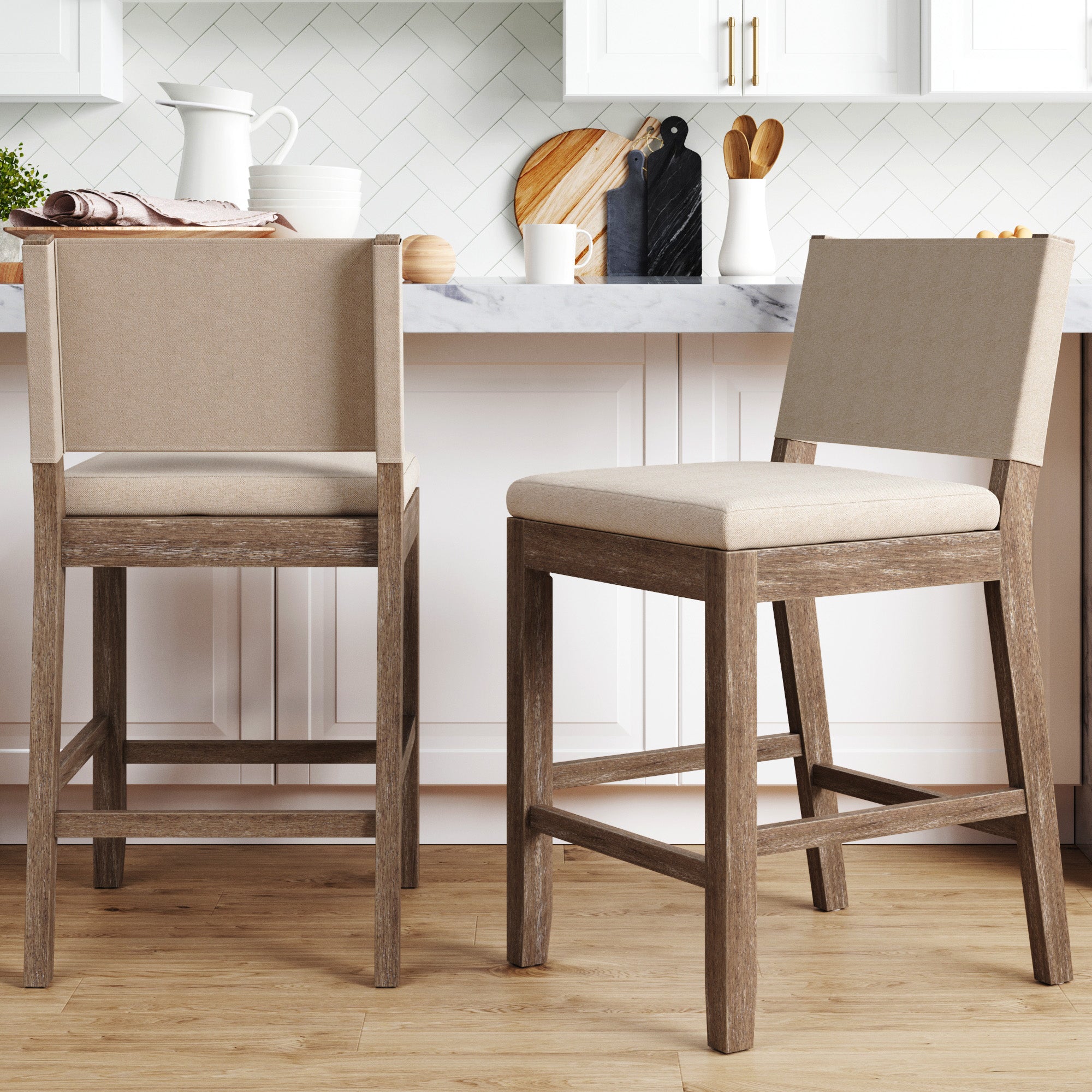 Two beige upholstered bar stools with wooden legs are placed in front of a kitchen counter with a marble top; the kitchen features white cabinets, a wood floor, and various kitchen items on the counter.