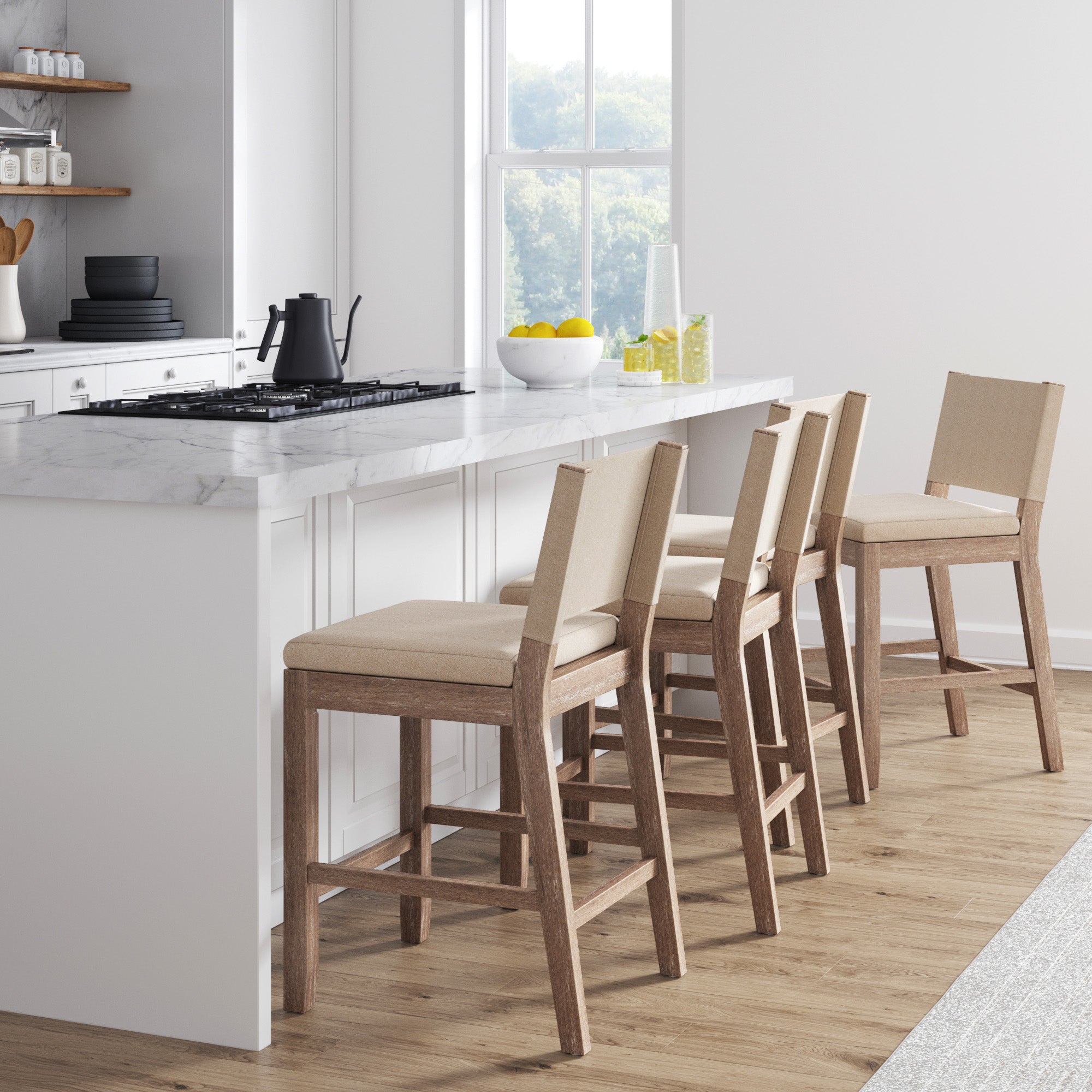A modern kitchen with a white marble island, four beige bar stools with wooden legs, a bowl of lemons, and sunlight coming through a window onto the wooden floor.