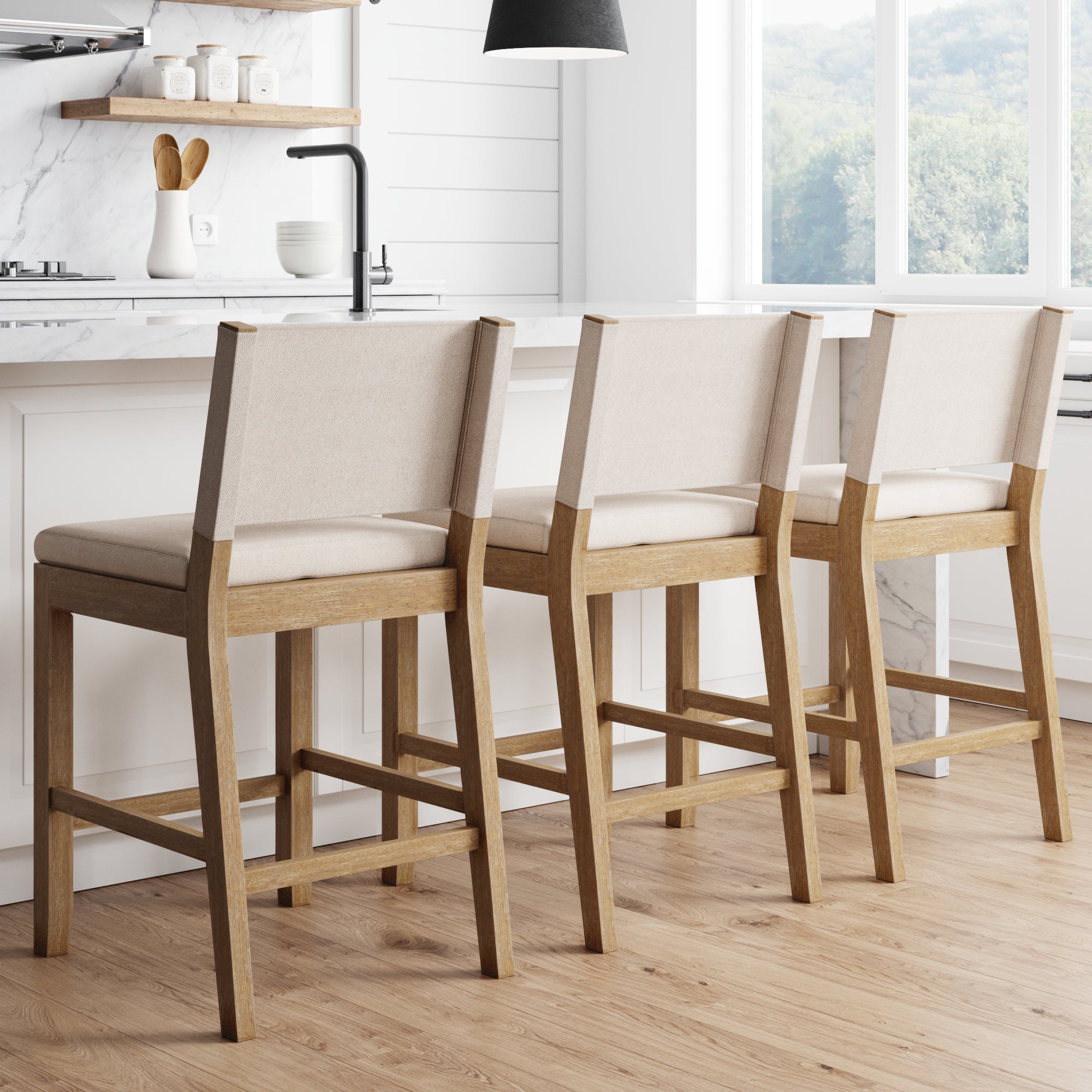 Three Nathan James Wood Counter Height Bar Stools in light brown with flax fabric seats and backs are lined up at a white kitchen island in a bright, modern farmhouse kitchen with light wood floors and large windows.