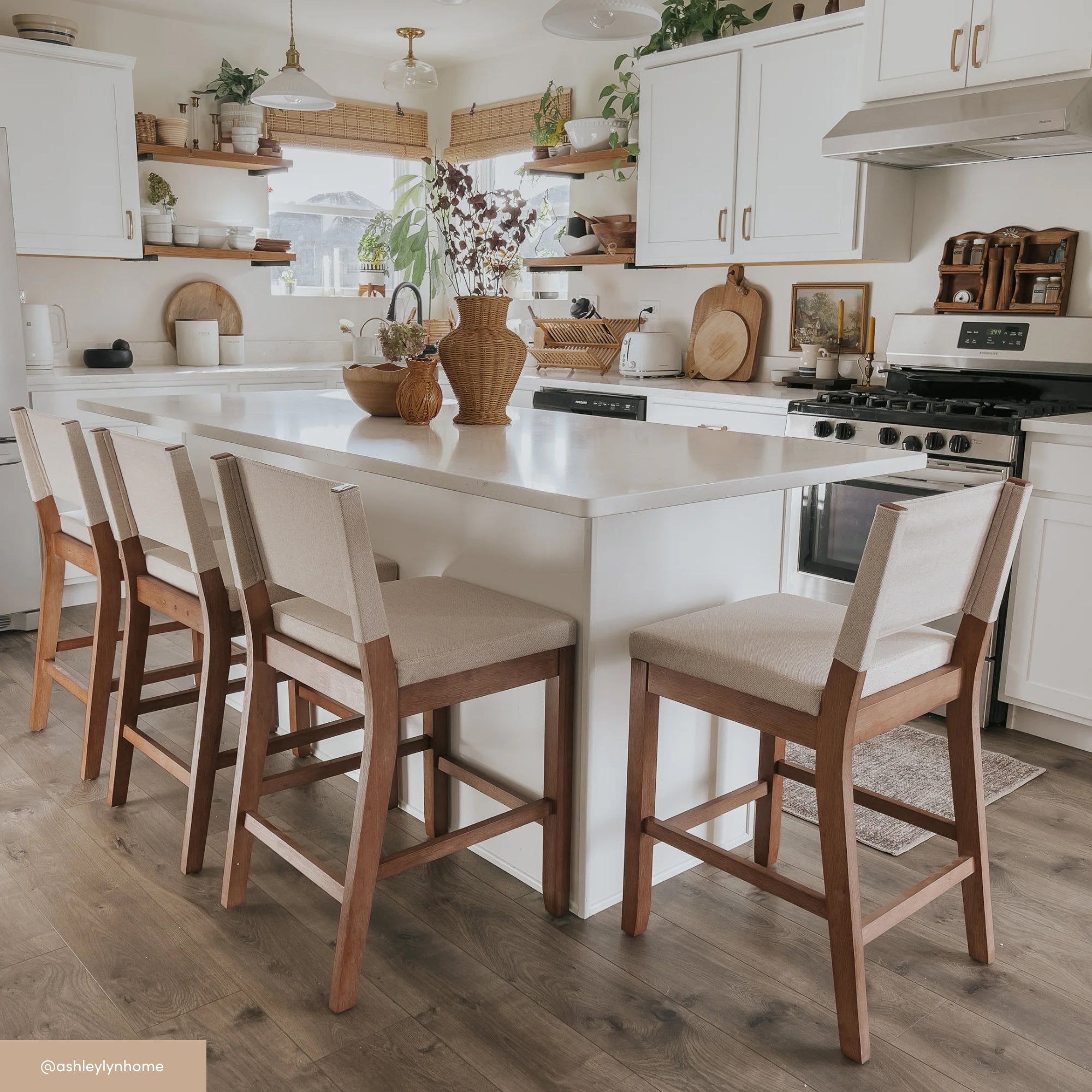 A bright, modern kitchen with white cabinets, a large island with four cushioned chairs, wooden accents, potted plants, and natural light streaming through windows with bamboo blinds.