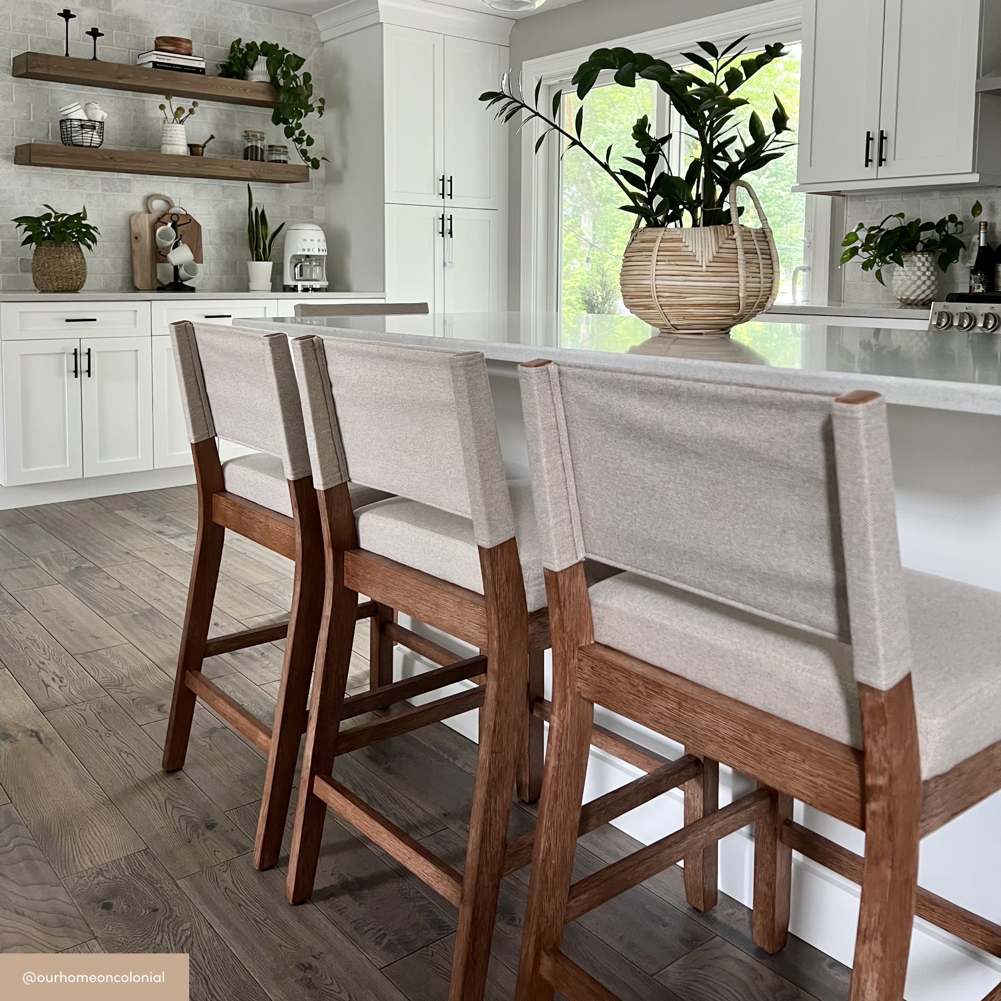 Modern kitchen with white cabinets, a white island with three wooden barstools, light wood floors, open shelves with decor, and large plants adding a fresh touch. Natural light streams in through a window.
