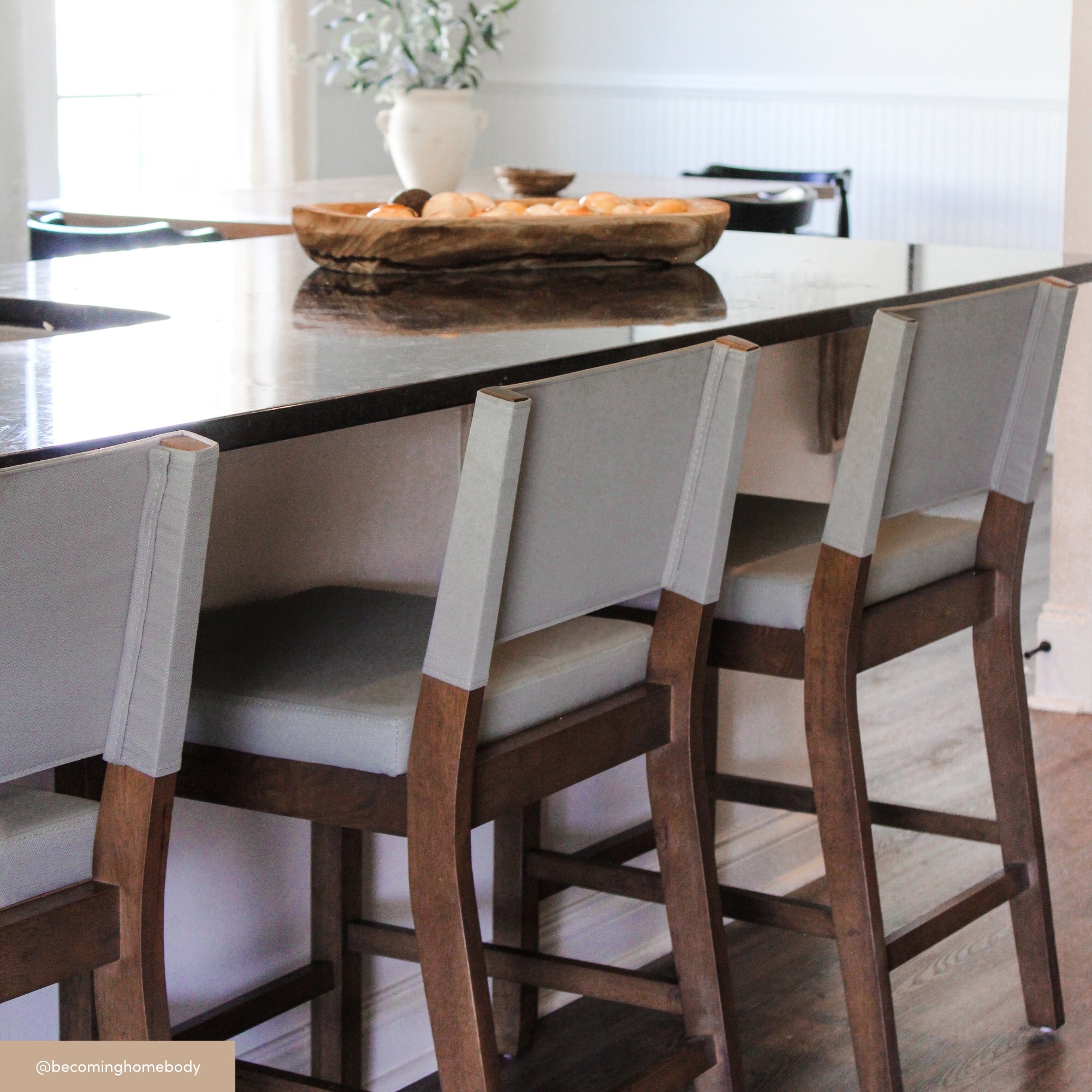 Modern kitchen with a dark stone countertop island, three wooden barstools with light gray cushions and backs, a wooden tray with oranges, and a white vase with greenery in the background.