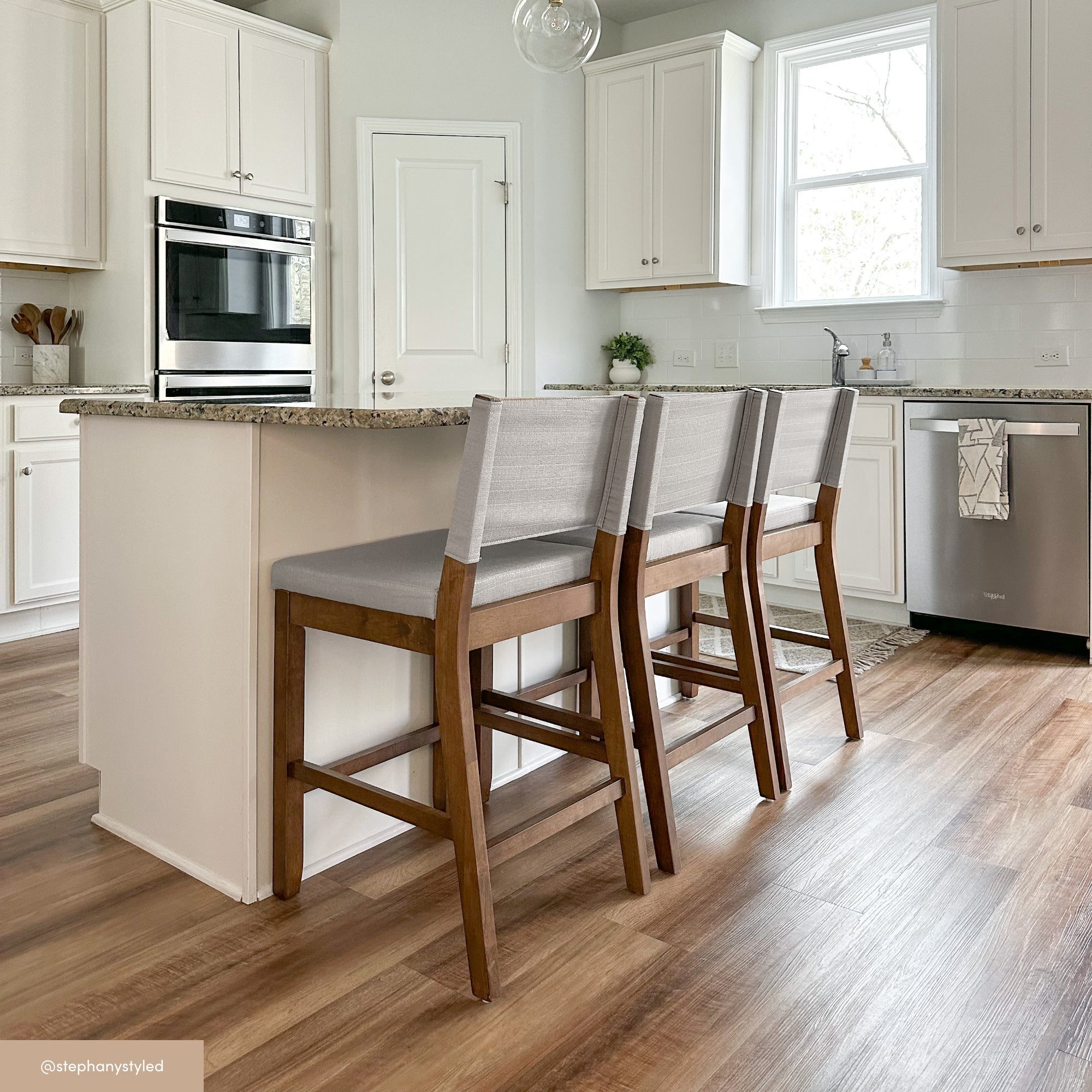 A modern kitchen with white cabinets, stainless steel appliances, and a granite island. Three wooden bar stools with light gray cushions are lined up at the island on a wood floor. Natural light enters through a window.