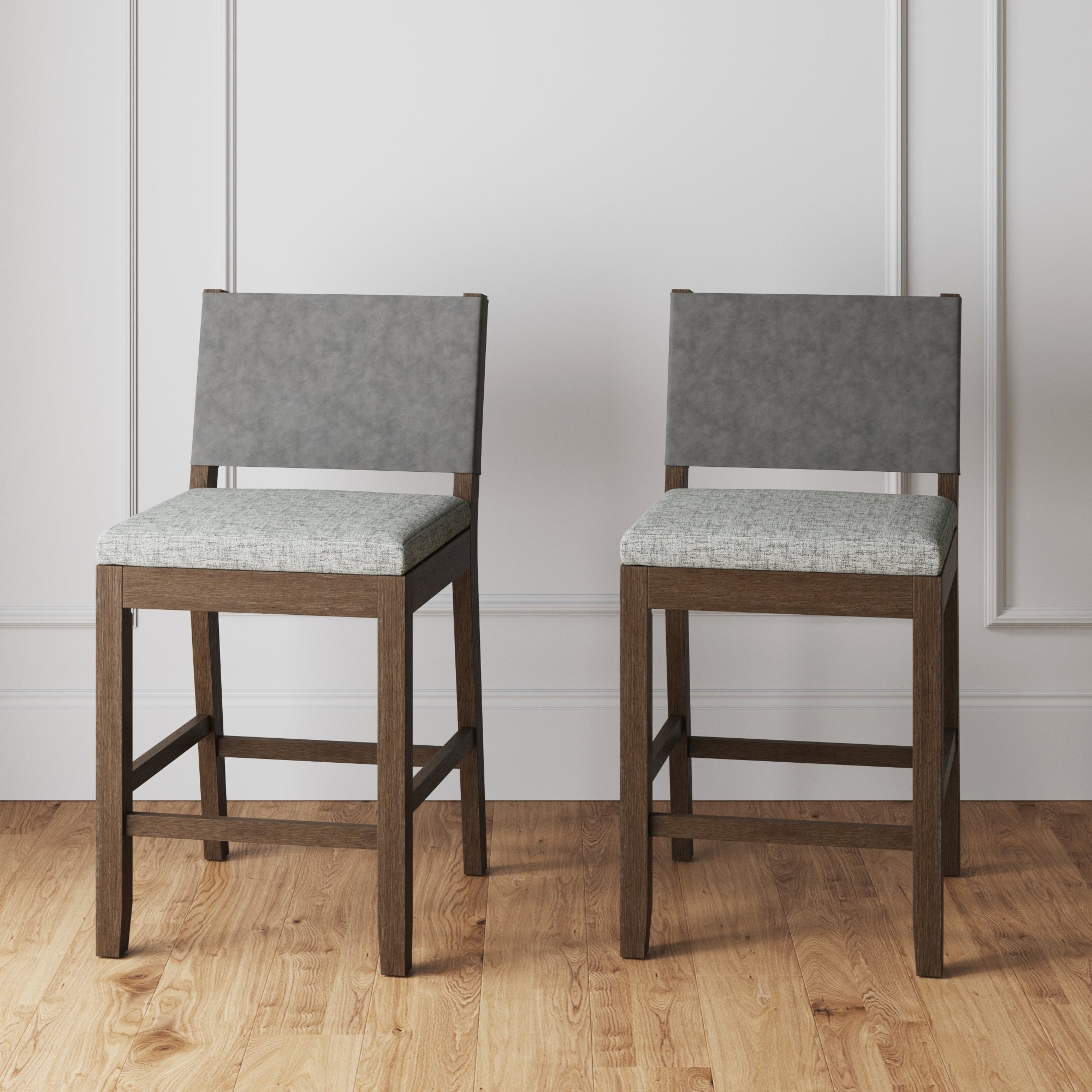Two modern bar stools with dark wooden legs and frames, light gray upholstered seats, and matching gray backrests, placed on a light wooden floor against a white paneled wall.