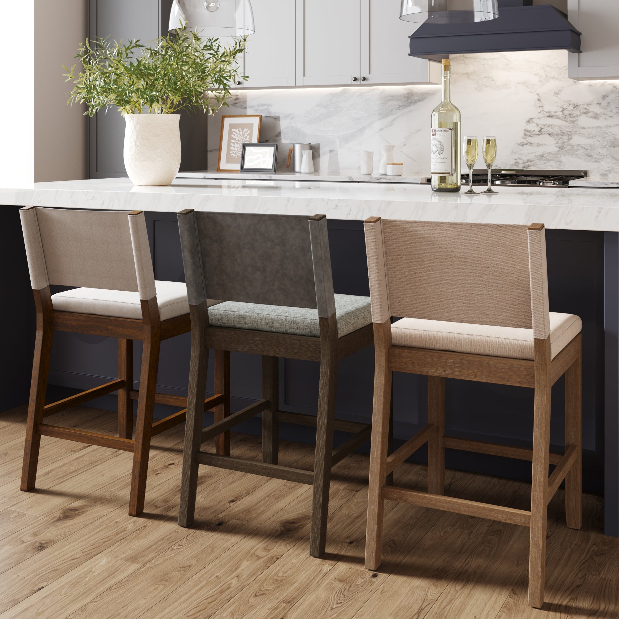 Three wooden barstools with fabric backs and seats in beige, gray, and tan are lined up at a marble kitchen island. A white vase with greenery, wine bottle, two glasses, and framed art sit on the countertop.