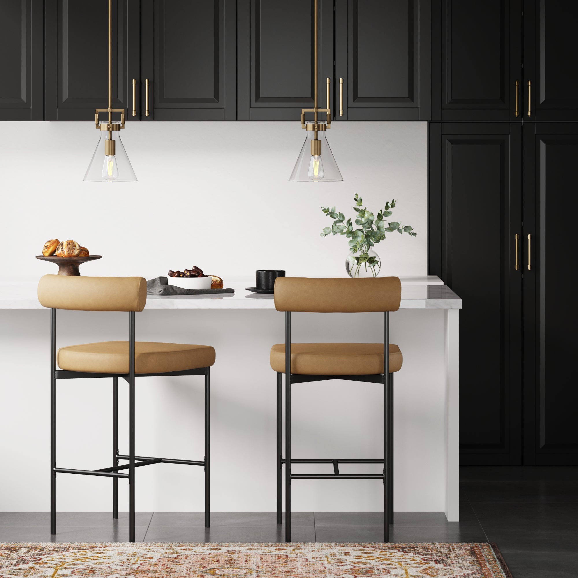A modern kitchen with black cabinets, gold handles, two tan barstools at a white island, pendant lights, pastries on a cake stand, a small plant, and a decorative rug visible at the bottom.