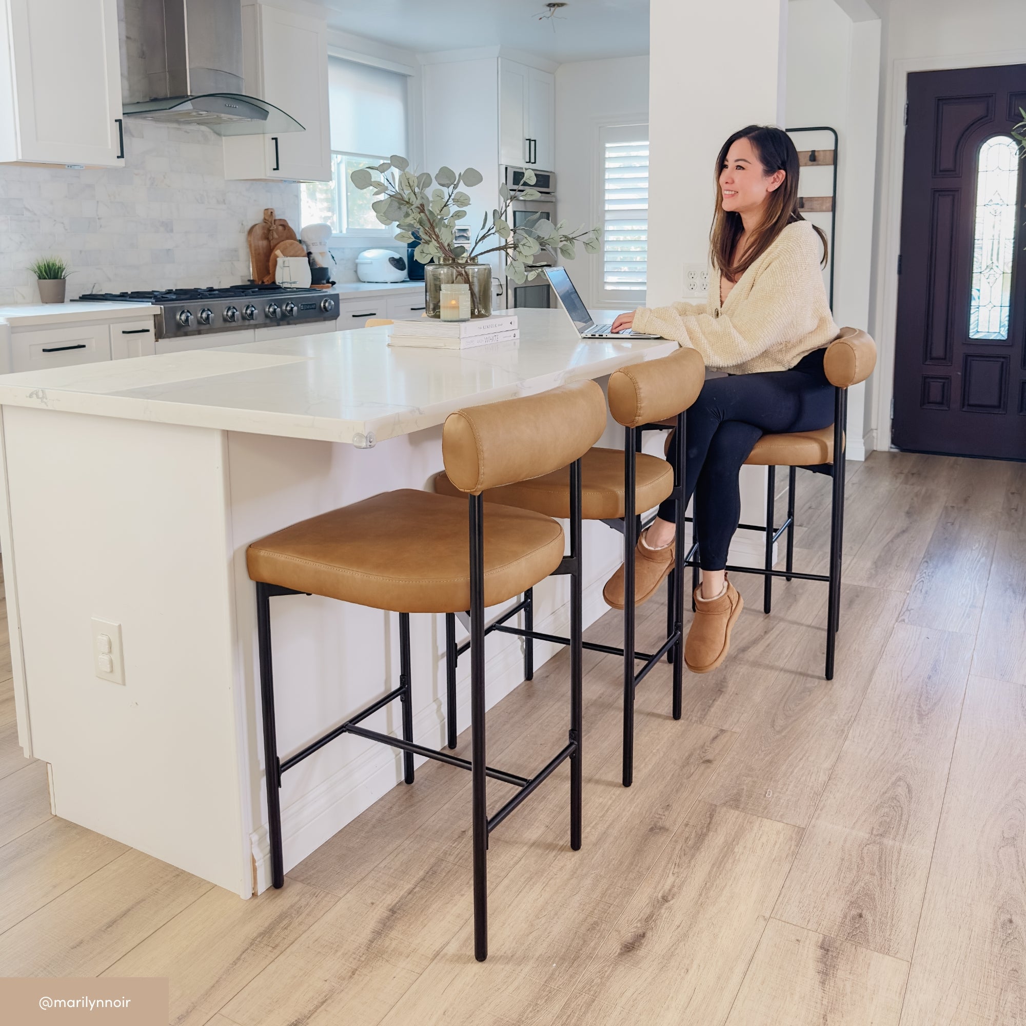 A woman sits on a tan barstool at a white kitchen island, working on a laptop. The kitchen is modern with white cabinets, wood floors, and minimalist decor. Natural light fills the space.