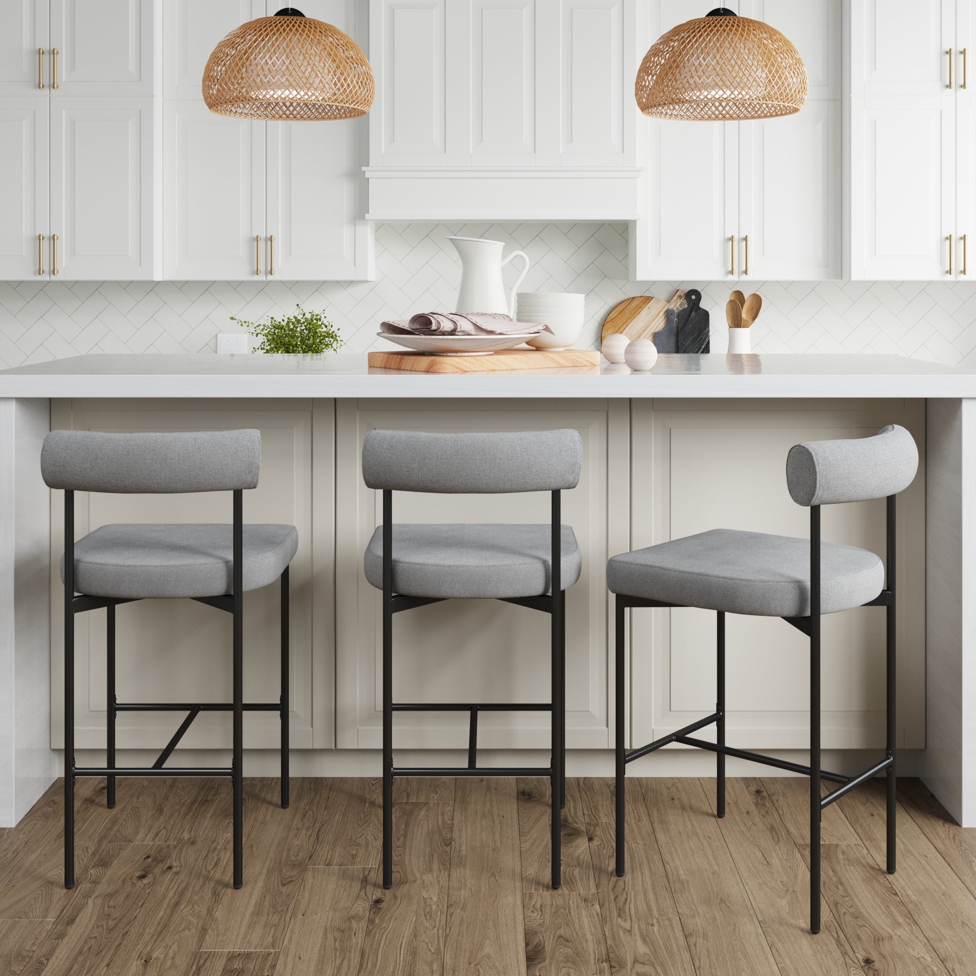 A modern kitchen with white cabinets, a white countertop island, and three Nathan James Metal Counter Height Bar Stools in Gray. Wooden cutting boards, kitchenware, and two woven pendant lights hang above.