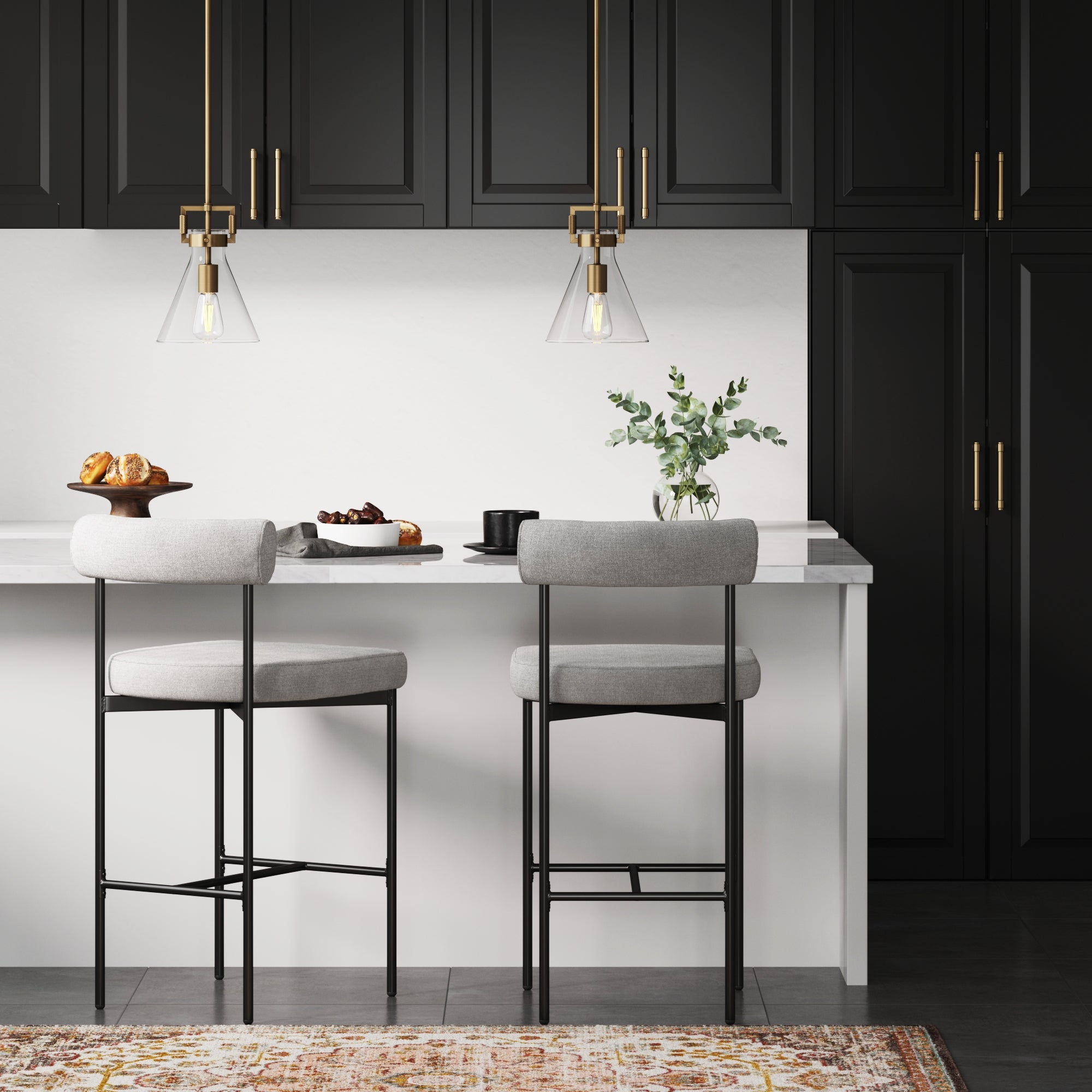 Modern kitchen with black cabinets, brass hardware, two pendant lights, a white island with two gray cushioned barstools, pastries on the counter, a black mug, and a vase with green foliage.