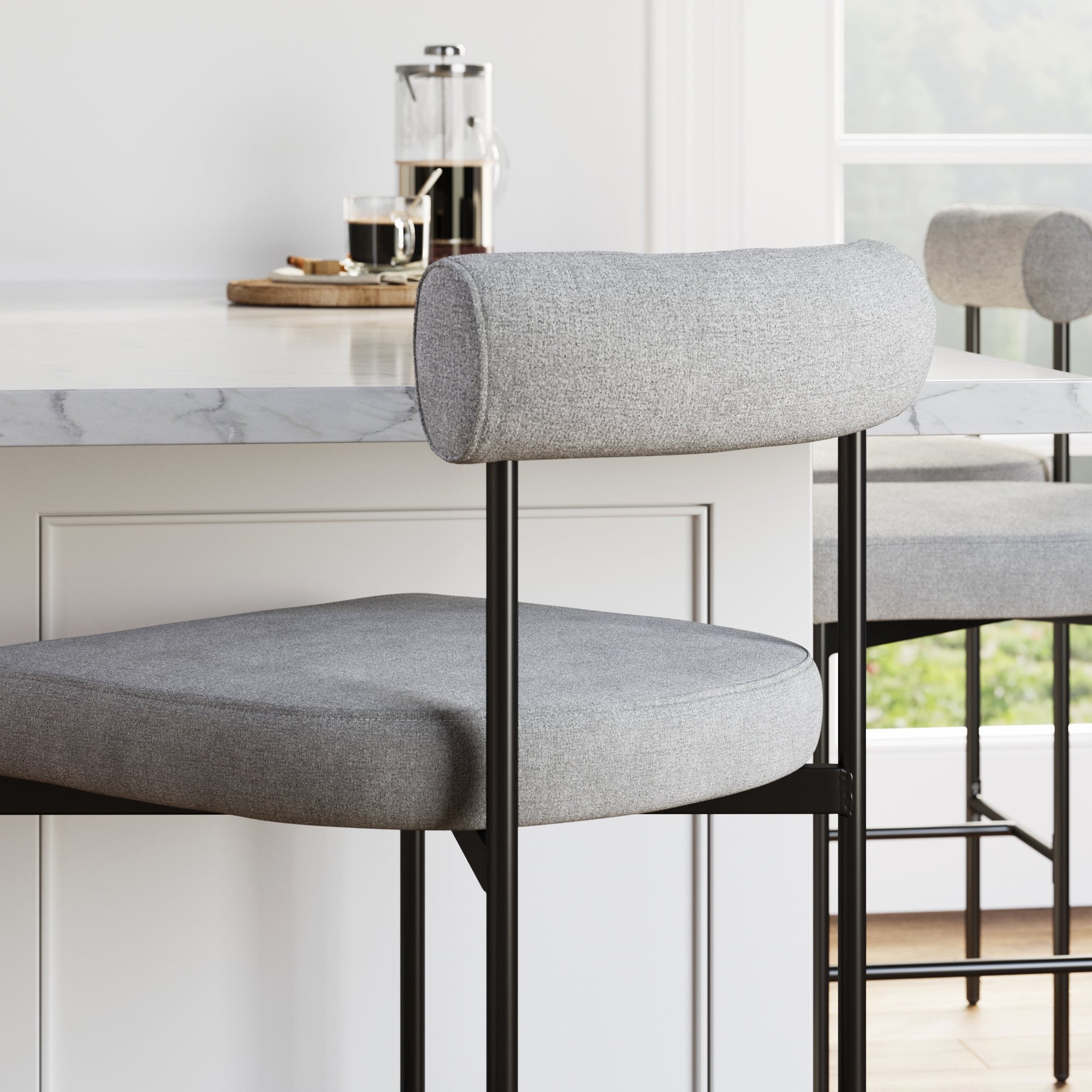 Modern kitchen with a marble countertop, a gray upholstered barstool with a metal frame, and a French press coffee maker with cups on a wooden tray in the background by a window.