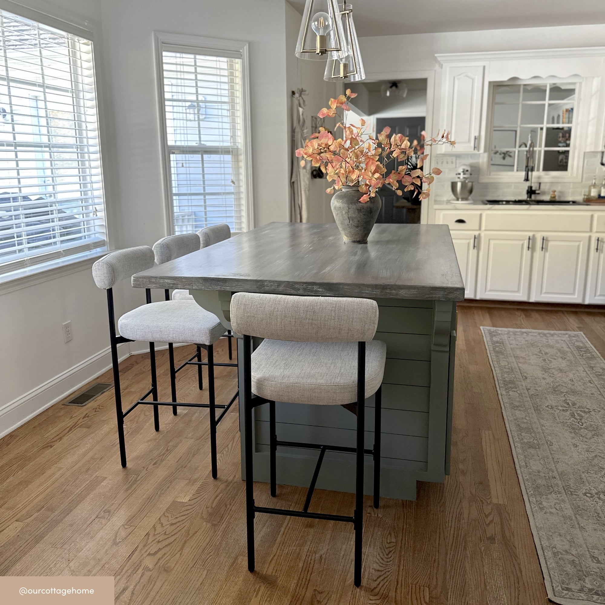 A modern kitchen with wooden floors, a gray island with two light gray cushioned barstools, a vase of peach flowers on the island, white cabinets, and large windows letting in natural light.