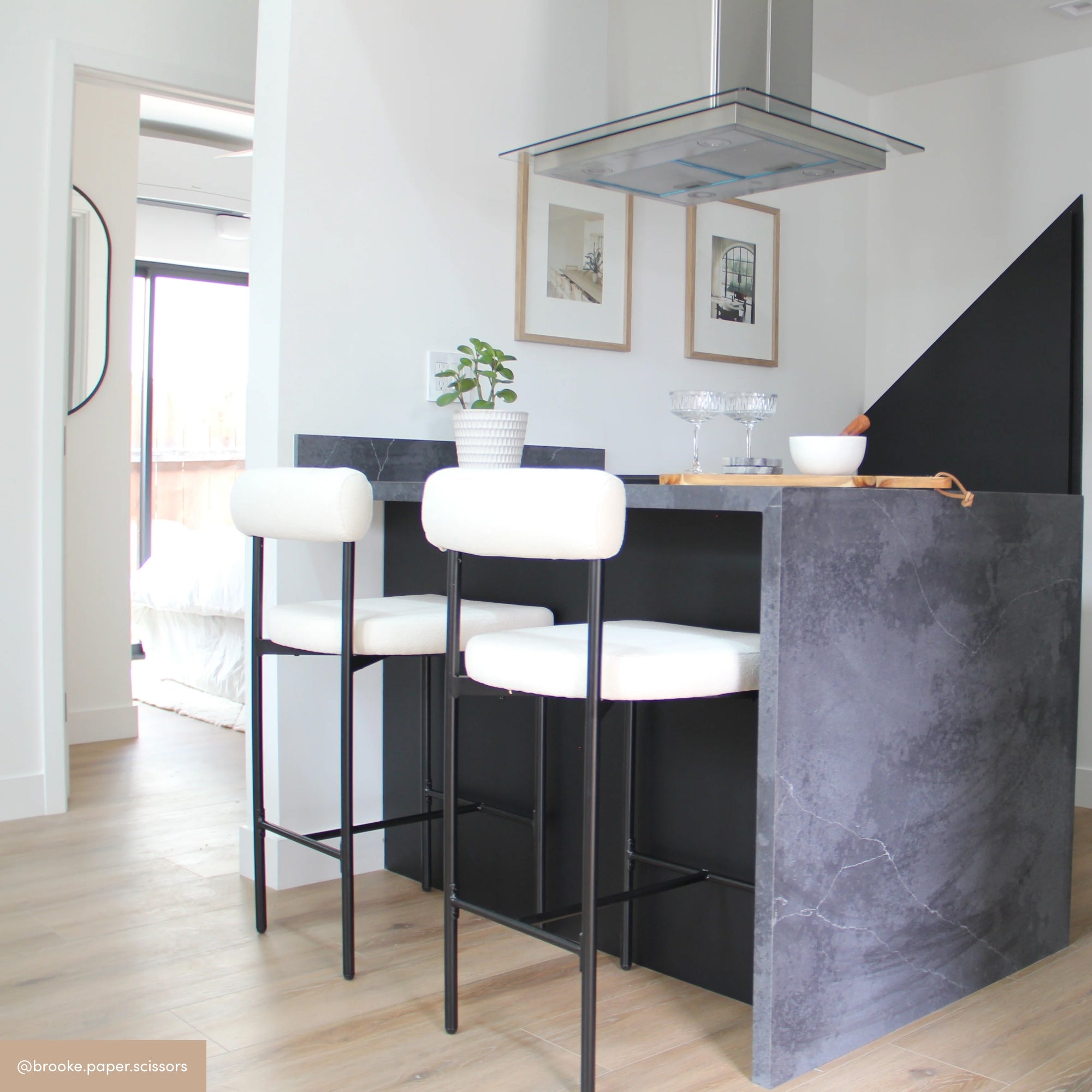 A modern kitchen with a gray marble island, two white barstools, a potted plant, glasses, and framed art on white walls. Light wood flooring and a doorway lead to a bright bedroom in the background.