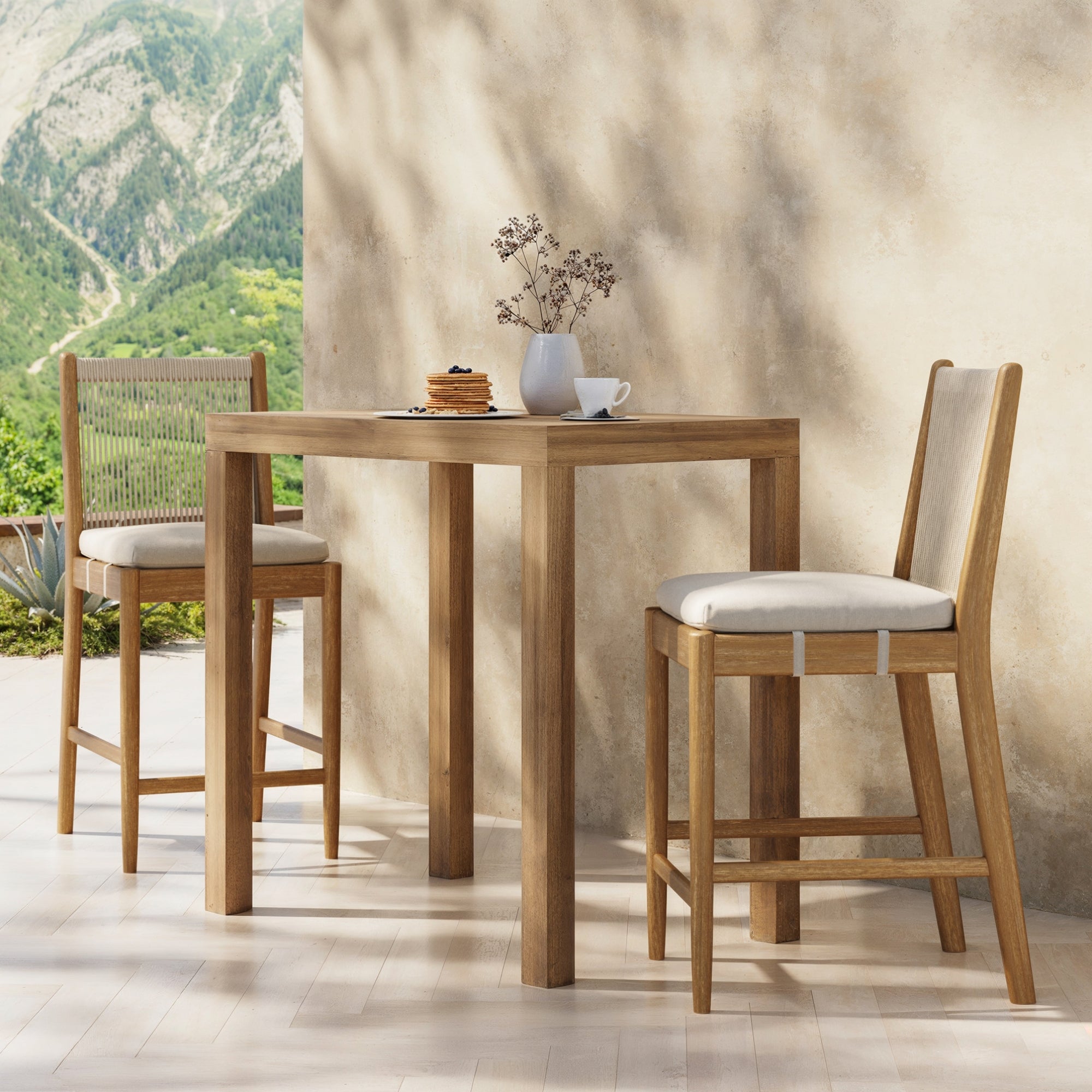 On a patio, Nathan James Outdoor Wood & Rope Counter Height Bar Stools (set of 2) in brushed light brown are paired with a wooden table, topped with a cup, pancakes, and vase. Lush green mountains form the backdrop.